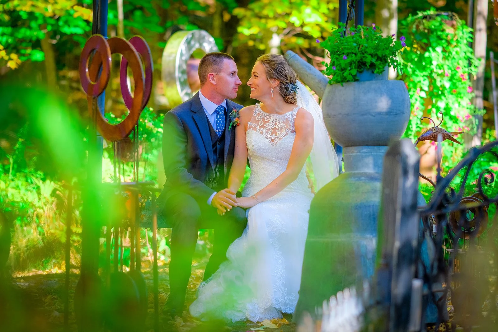A bride and groom sitting on a bench in a garden, gazing into each other's eyes with a backdrop of trees, colorful plants, and garden decorations.