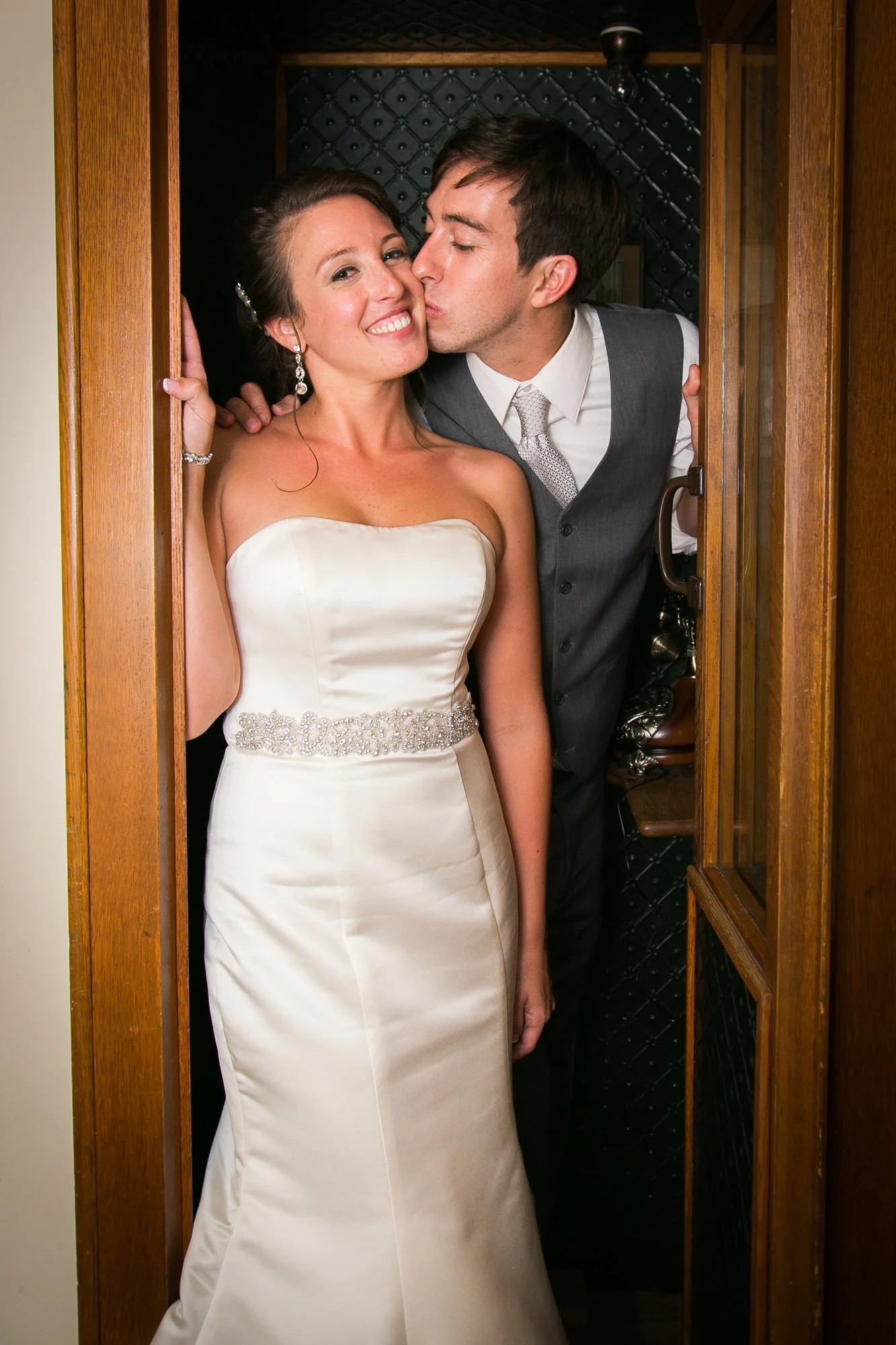 Bride and groom sharing a kiss inside a phone booth, with the groom about to kiss the bride on her cheek.