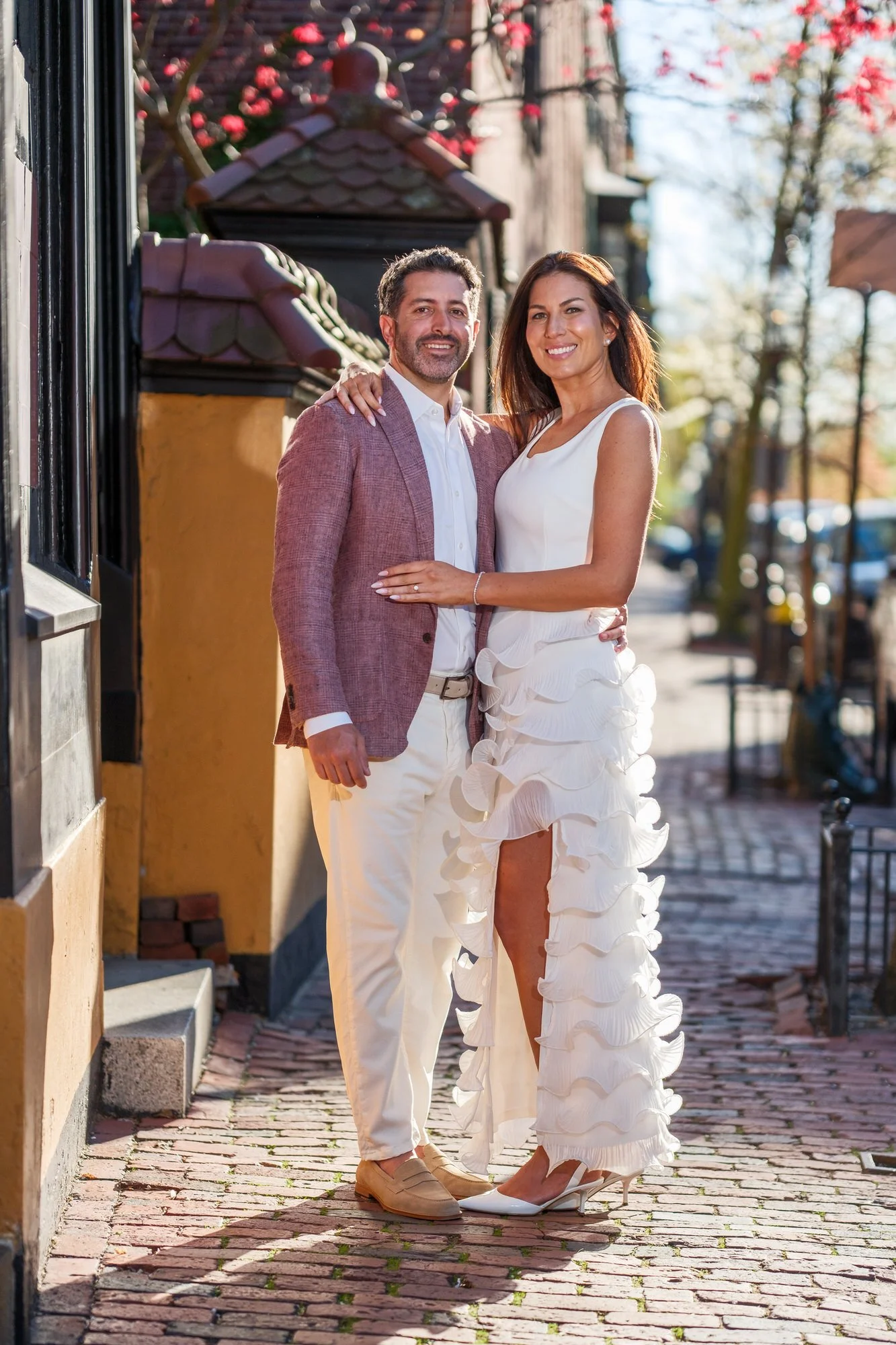 Lauren and Charlie posing together on a sun-drenched cobblestone sidewalk in Beacon Hill, Boston, arms around each other and smiling warmly at the camera with a red-tiled building behind them