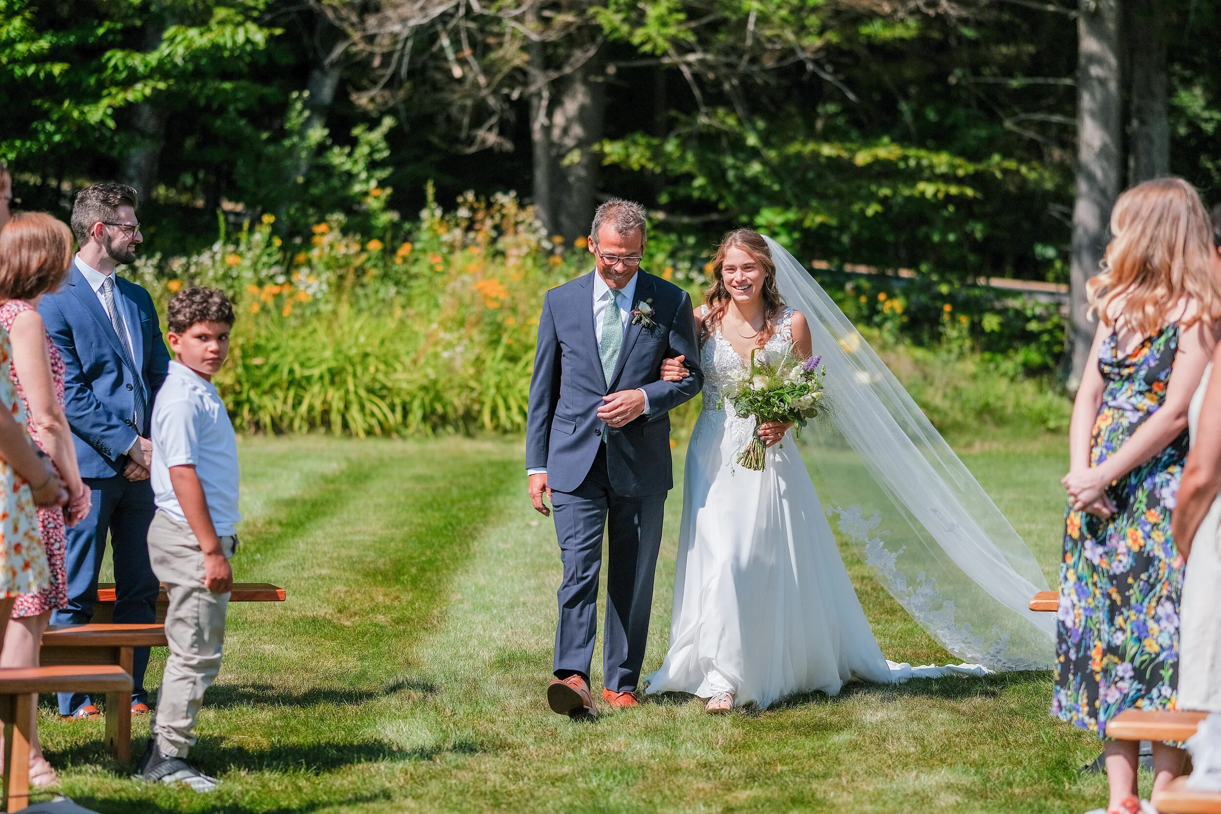 A bride walking down the outdoor wedding aisle with her father, holding a bouquet, surrounded by guests in formal attire, with a lush green garden background.