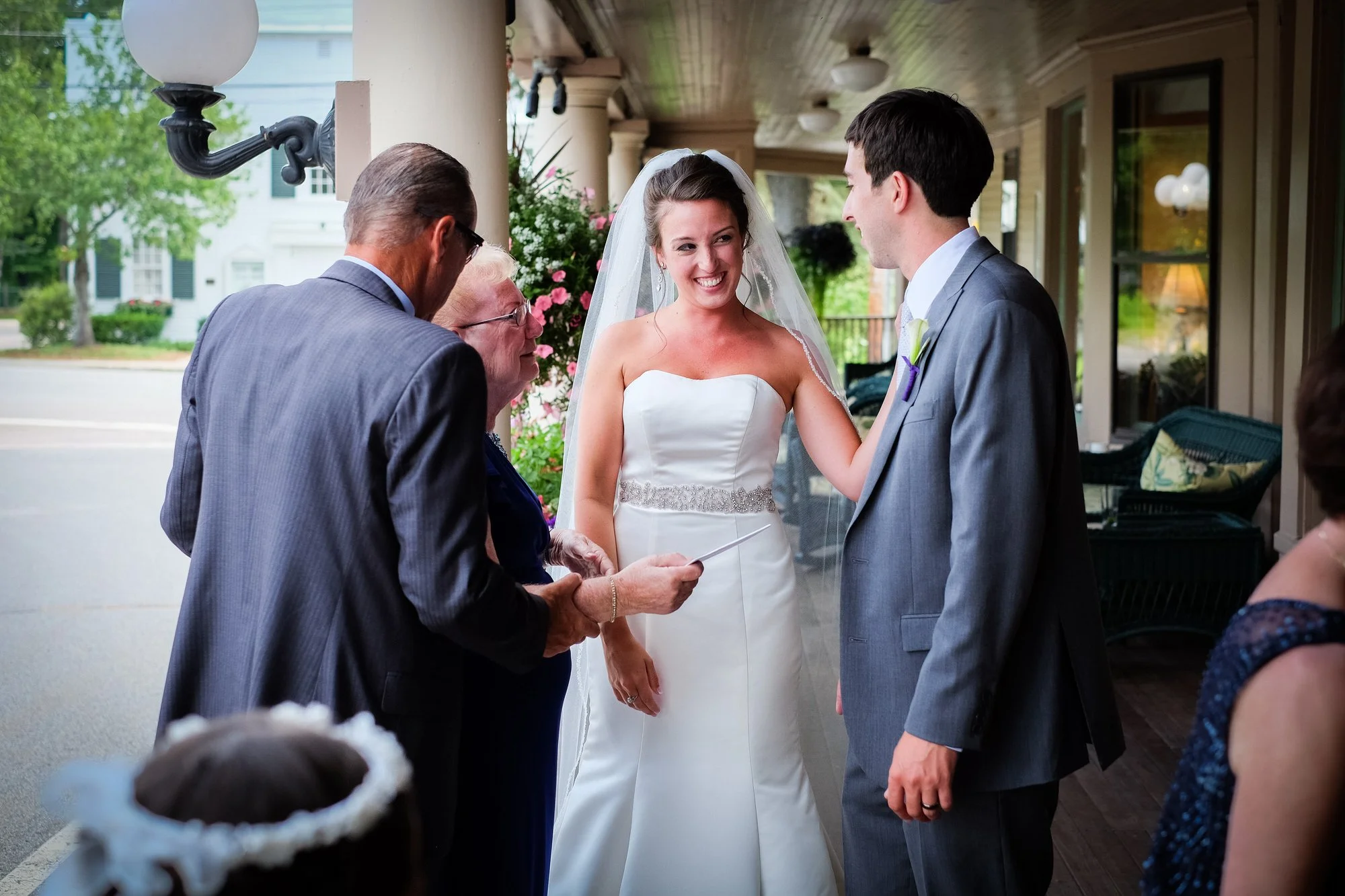 Bride and groom exchanging vows during wedding ceremony with officiant and guests on a porch.