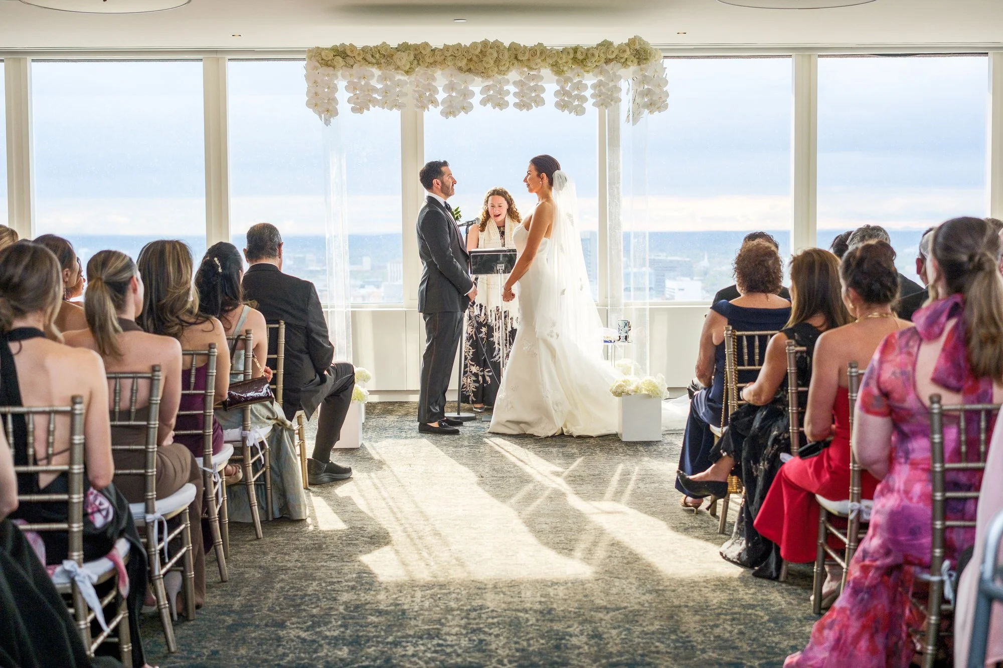 Wide ceremony shot of bride and groom under a white floral chuppah at the UMass Club Boston with panoramic city views