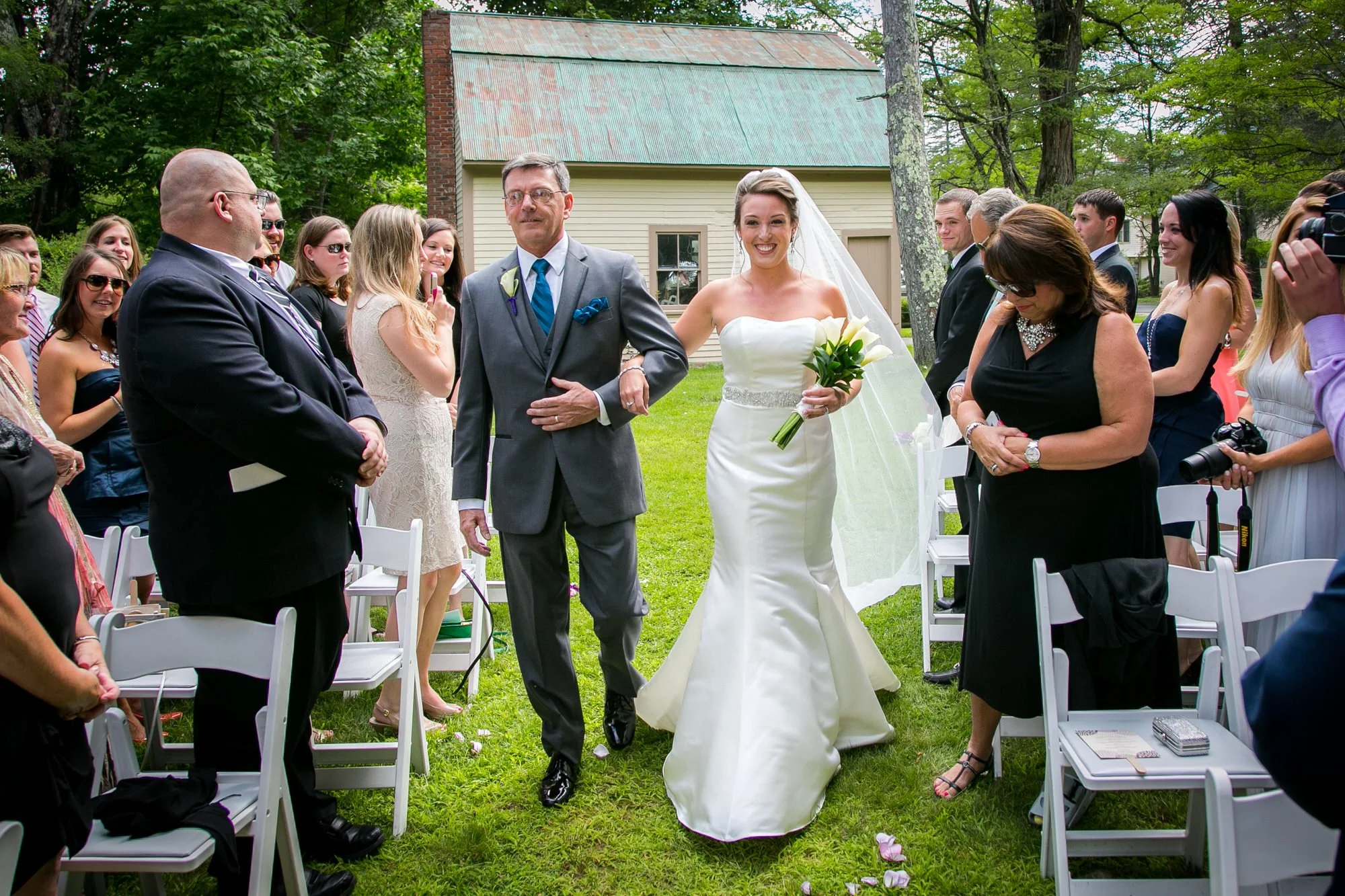 A bride walking down the aisle with her father at an outdoor wedding ceremony, surrounded by seated guests and greenery.