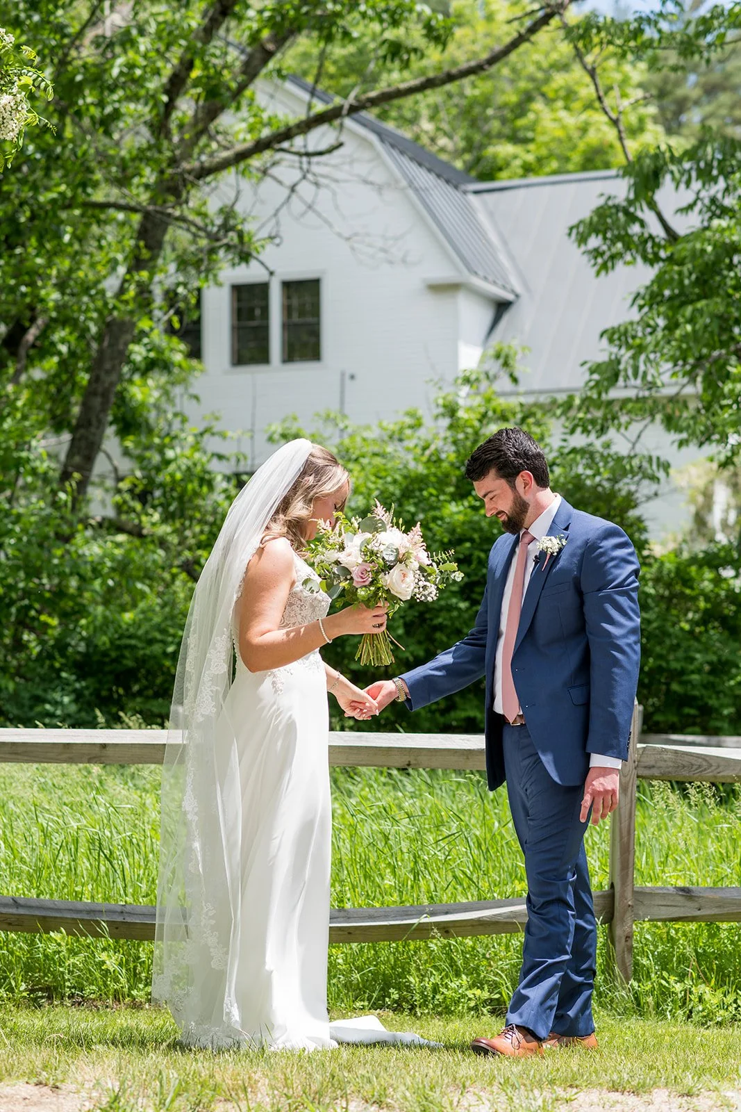 Bride and groom holding hands outdoors on a sunny day, with a white house and green trees in the background. The bride is wearing a white wedding dress and veil, and the groom is dressed in a blue suit. The bride holds a bouquet of flowers.