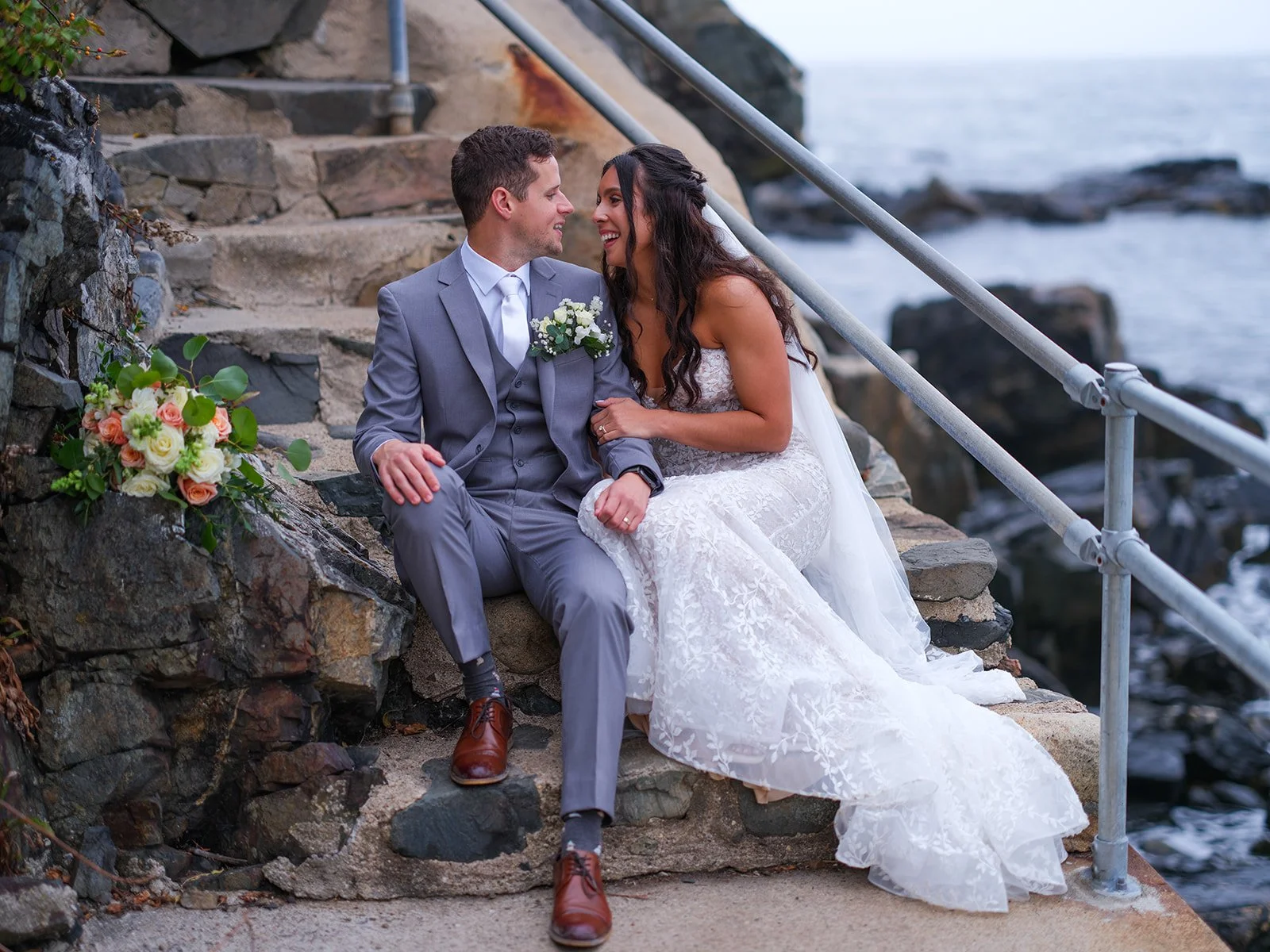 Bride and groom laughing together seated on stone steps by the ocean at York Maine fall wedding