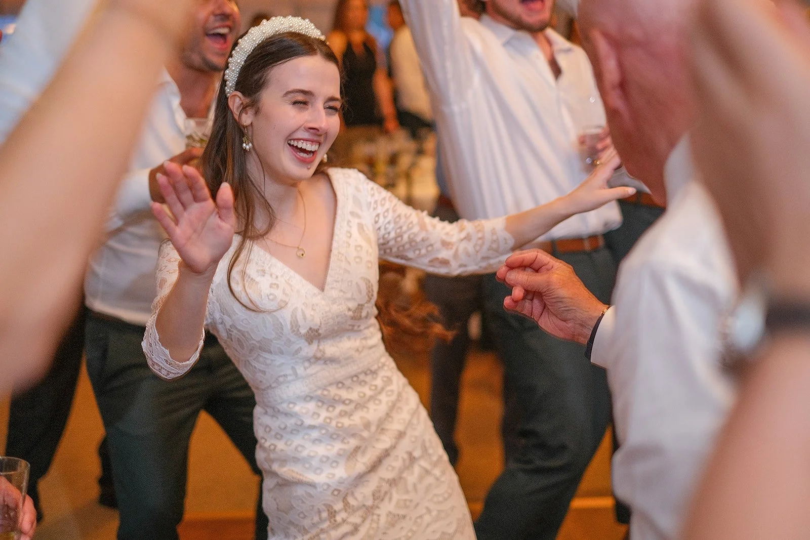 A young woman in a white lace dress and pearl headband is dancing and smiling with her eyes closed, while other people around her are also dancing and enjoying a celebration.