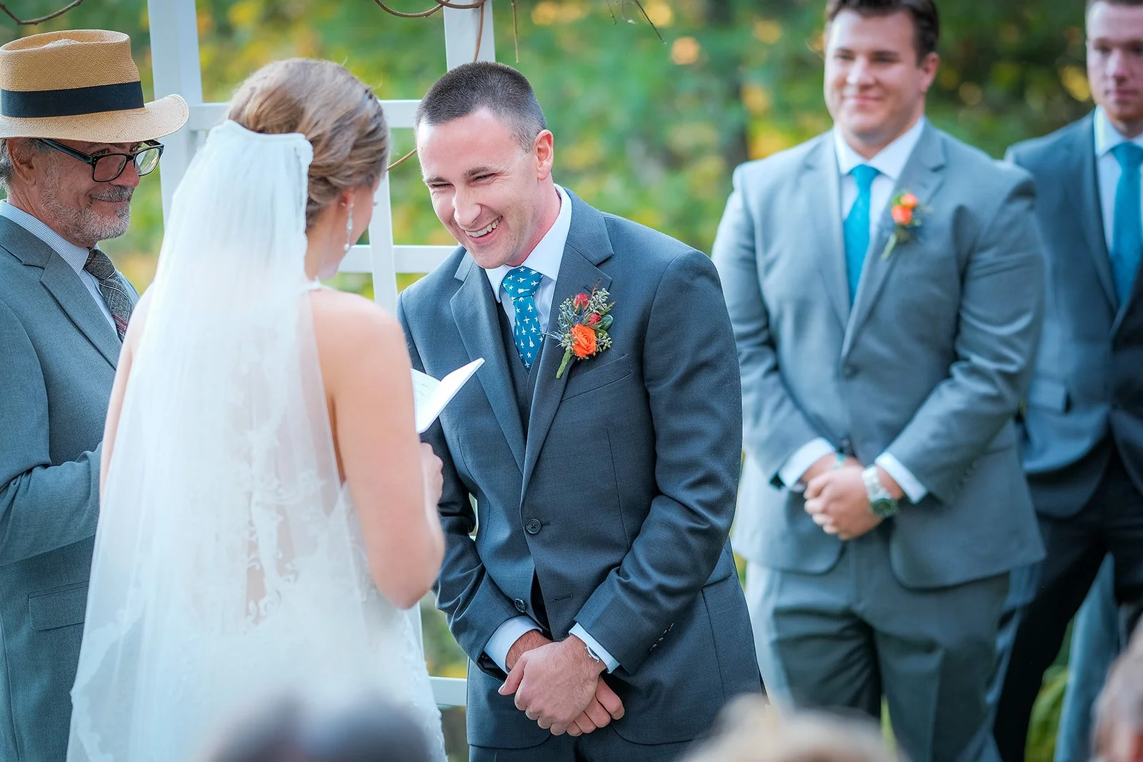 A bride and groom exchange vows during an outdoor wedding ceremony, with the bride reading from a paper and the groom smiling. Two groomsmen or family members stand nearby, dressed in suits, with a lush green background.