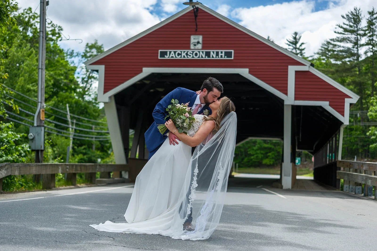 A newlywed couple shares a kiss on a street in front of a red covered bridge in Jackson, New Hampshire. The groom in a blue suit leans over the bride in a white wedding gown, holding a bouquet of flowers, with trees and a partly cloudy sky in the bac
