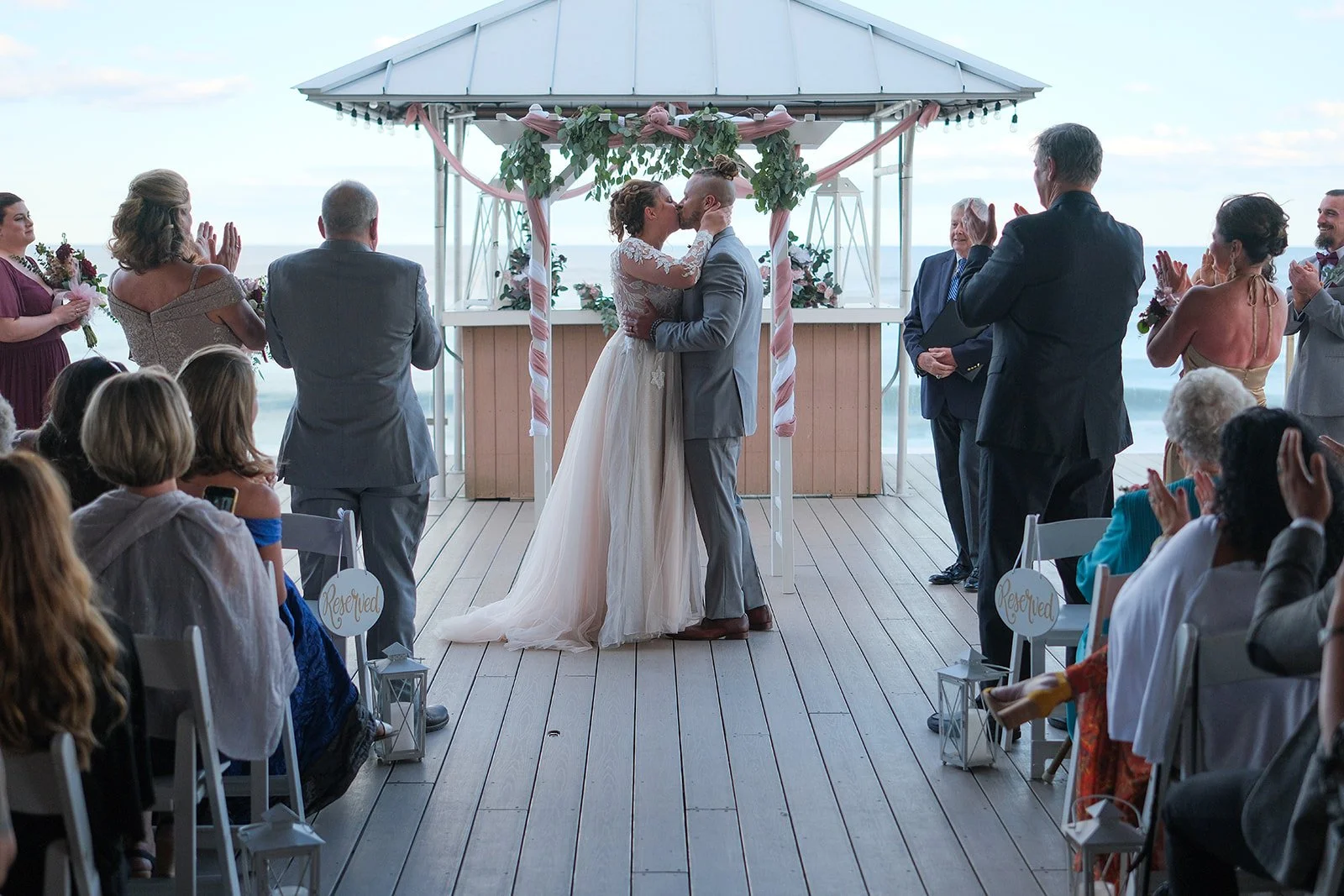 Bride and groom sharing first kiss on the oceanfront deck at Blue Ocean Event Center with guests celebrating around them