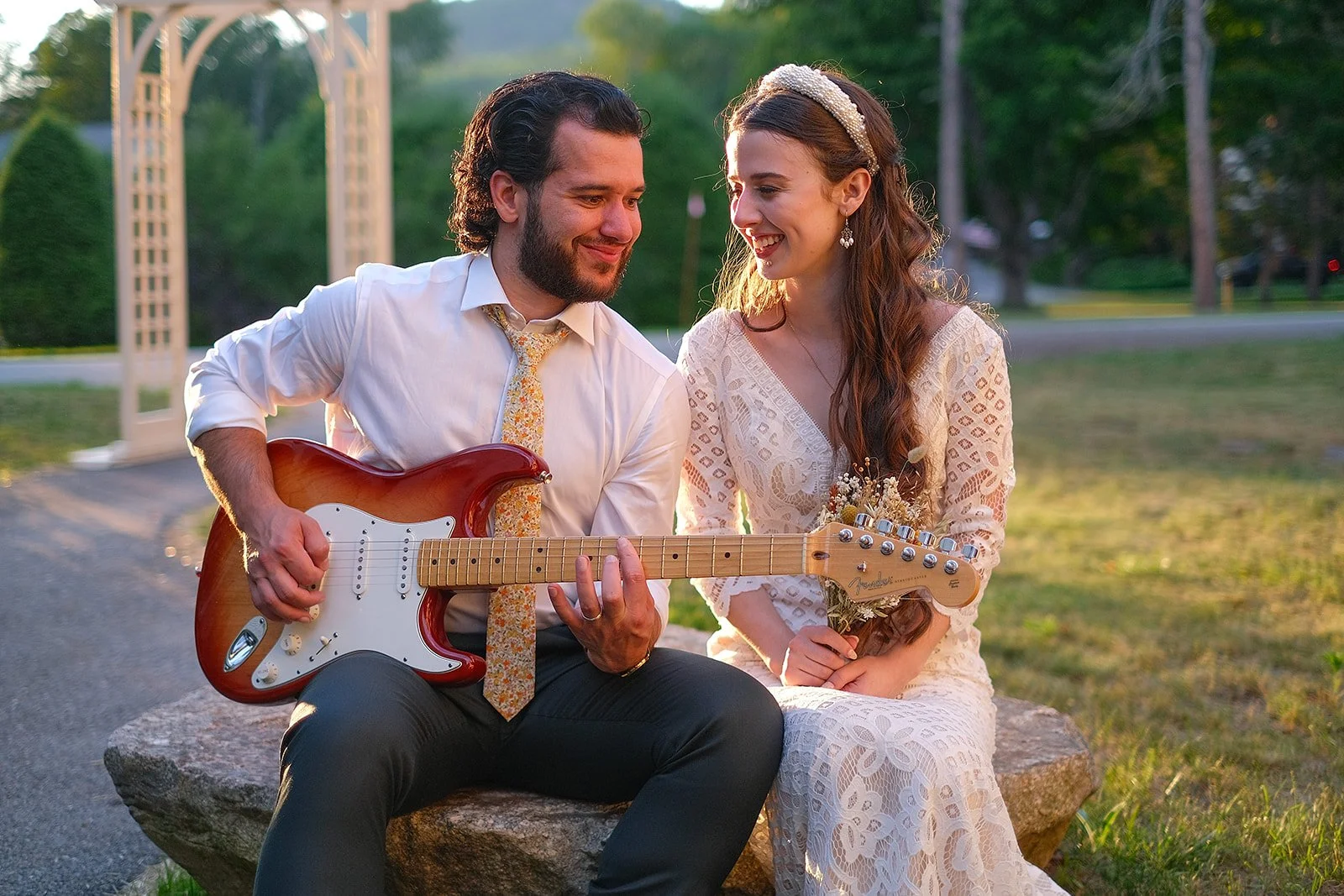 A man and woman sit on a rock outdoors during sunset, the man playing a guitar and both smiling at each other. The woman holds a small bouquet of dried flowers, and the man wears a white shirt with a patterned tie. In the background, there is a decor
