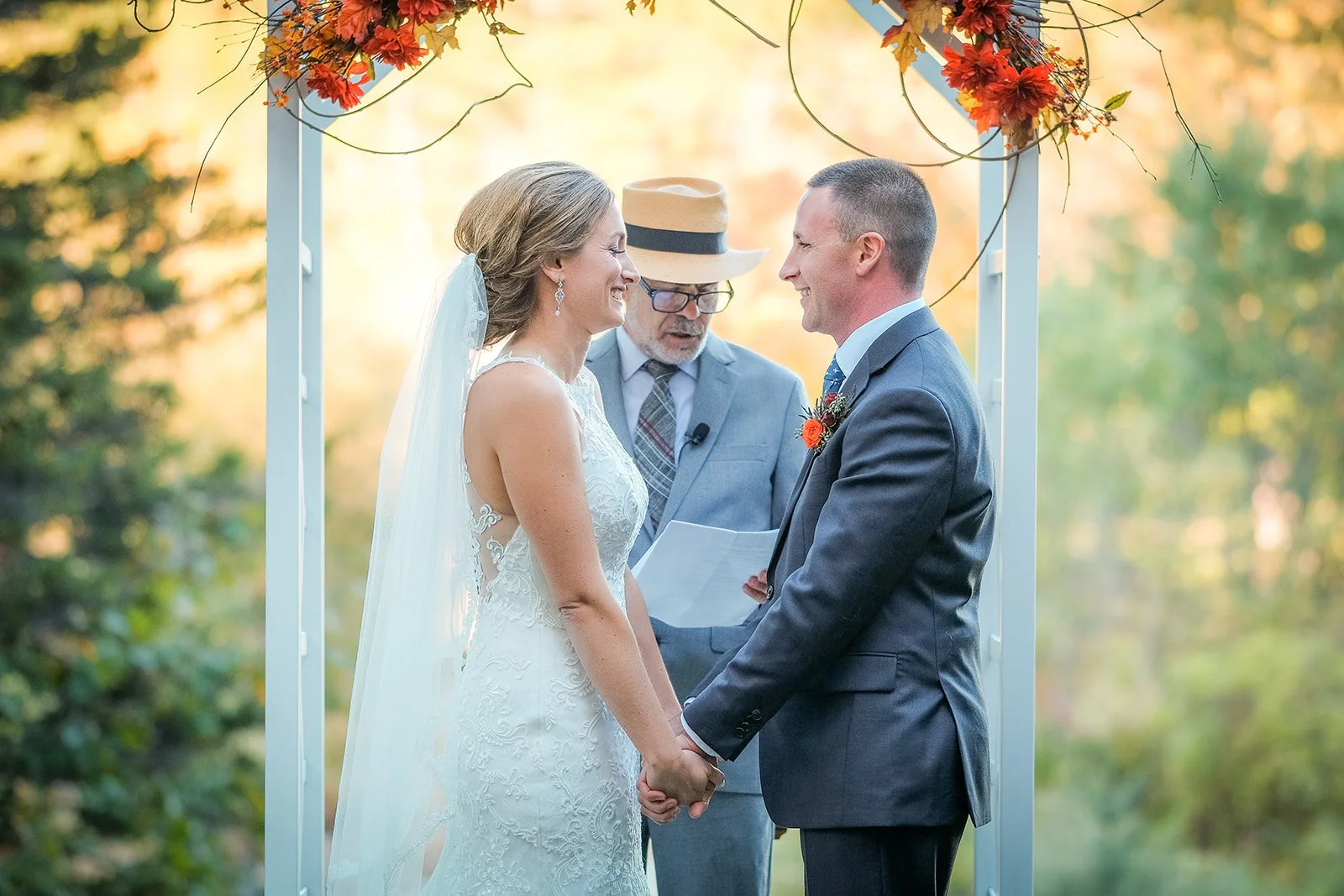 A bride and groom holding hands and smiling at each other during an outdoor wedding ceremony, with an officiant standing behind them and decorated with autumn-themed flowers and greenery.