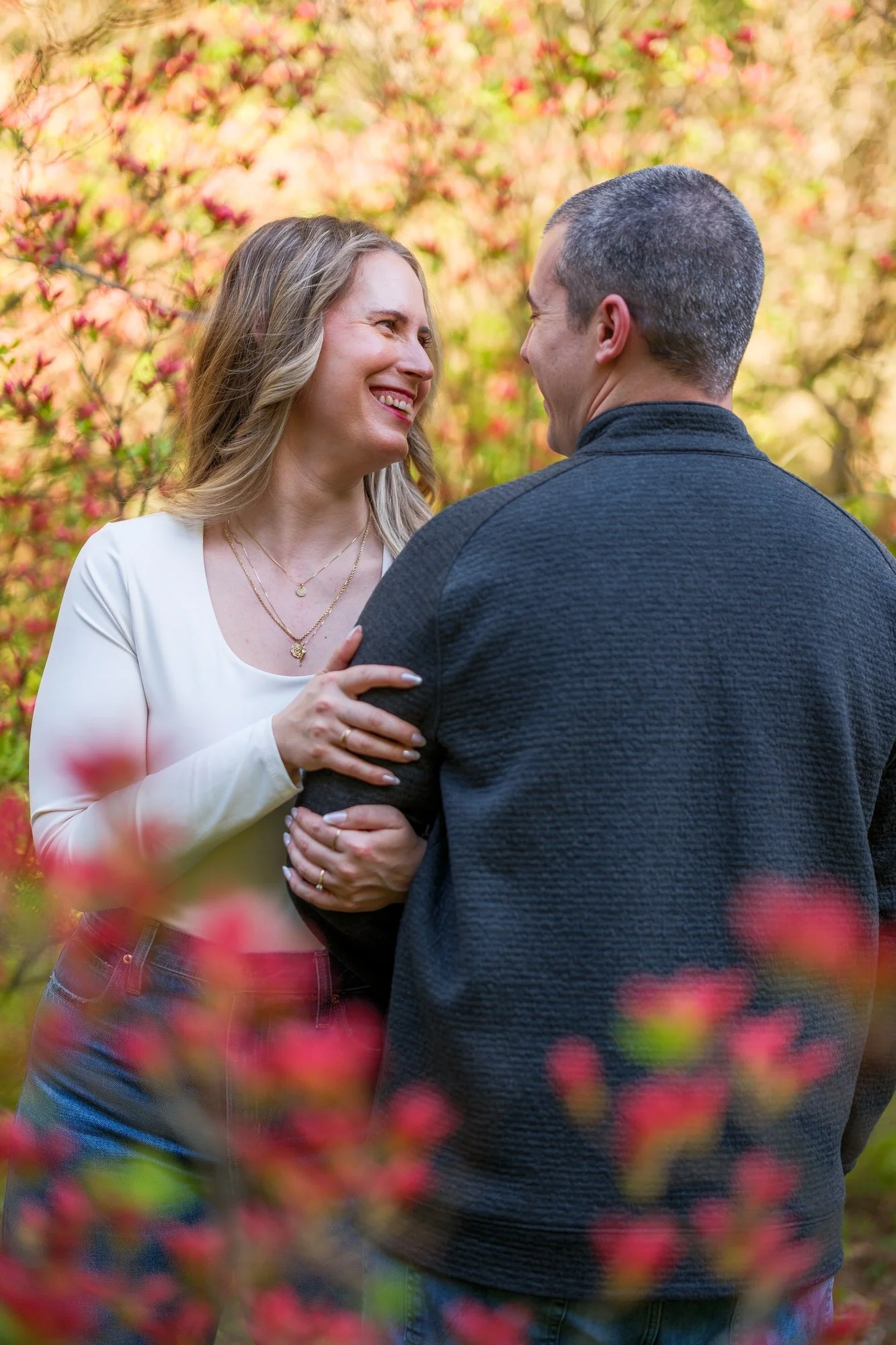 Candid portrait of bride-to-be laughing and holding her fiancé's arm surrounded by red flowering shrubs at Maudslay State Park