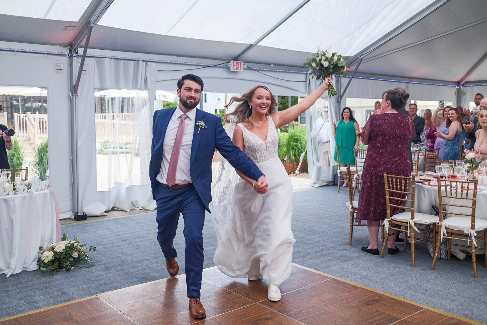 Bride and groom holding hands, walking onto dance floor at wedding reception, guests clapping and smiling in background, decorated tent with tables and chairs.