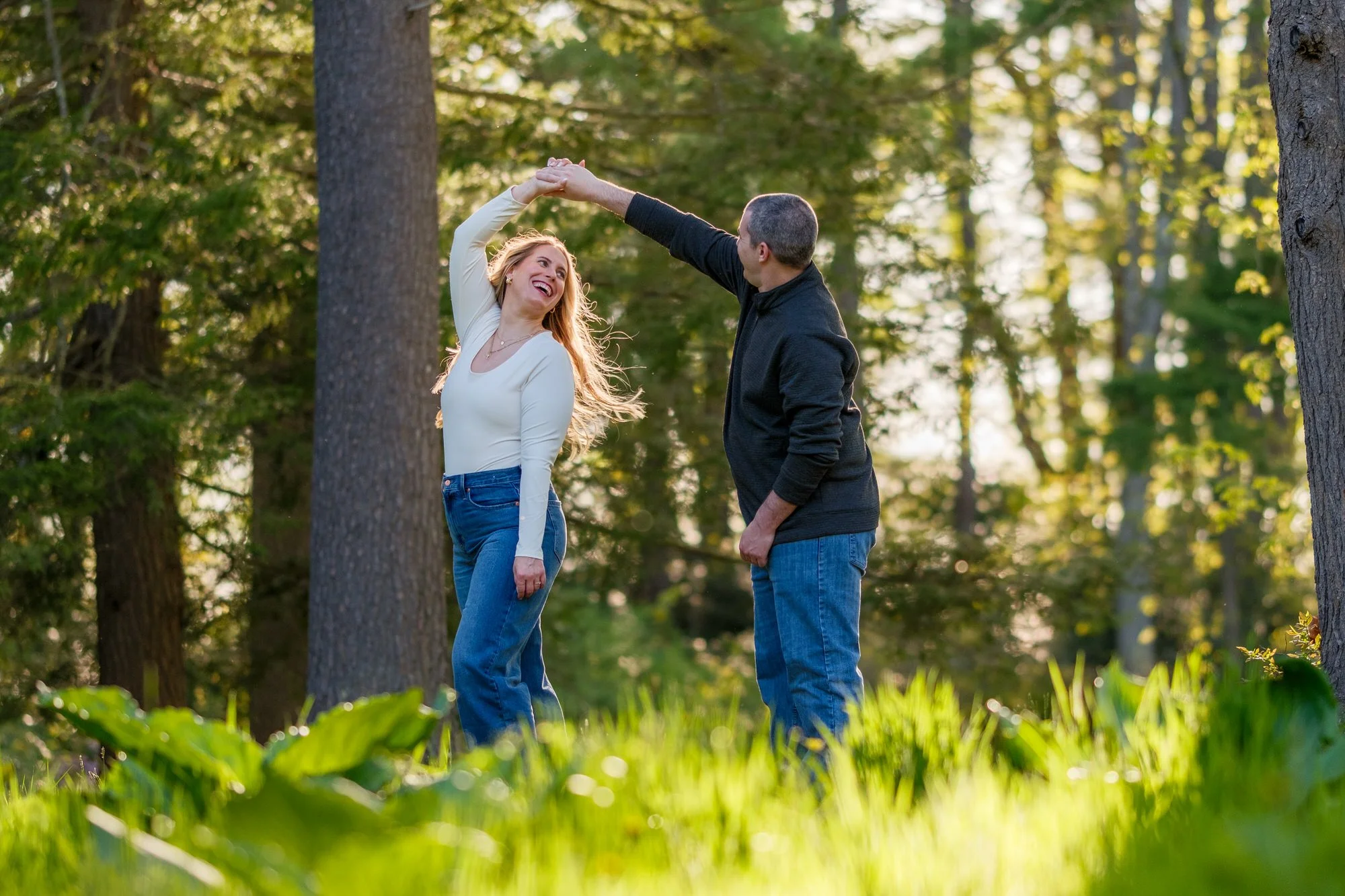 Groom spinning his fiancée in a sunlit pine forest meadow during their engagement session at Maudslay State Park