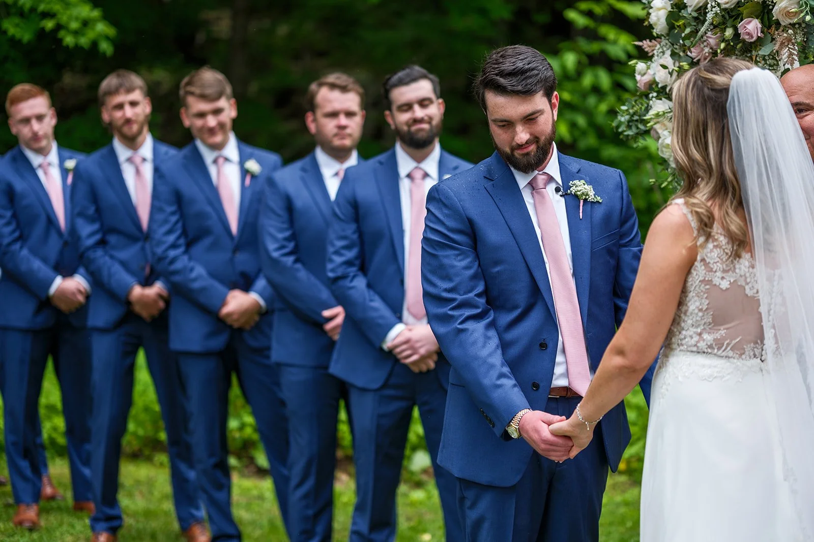 A groom and bride holding hands during a wedding ceremony, with the groom looking down and the bride partially visible wearing a veil. In the background, six groomsmen in blue suits and pink ties are standing in a line, outdoors with greenery.