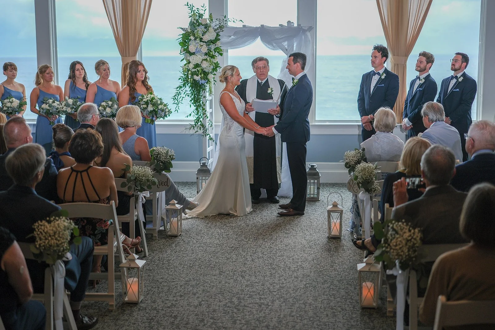 Wedding ceremony at Blue Ocean Event Center with ocean views through large windows and floral arch behind couple