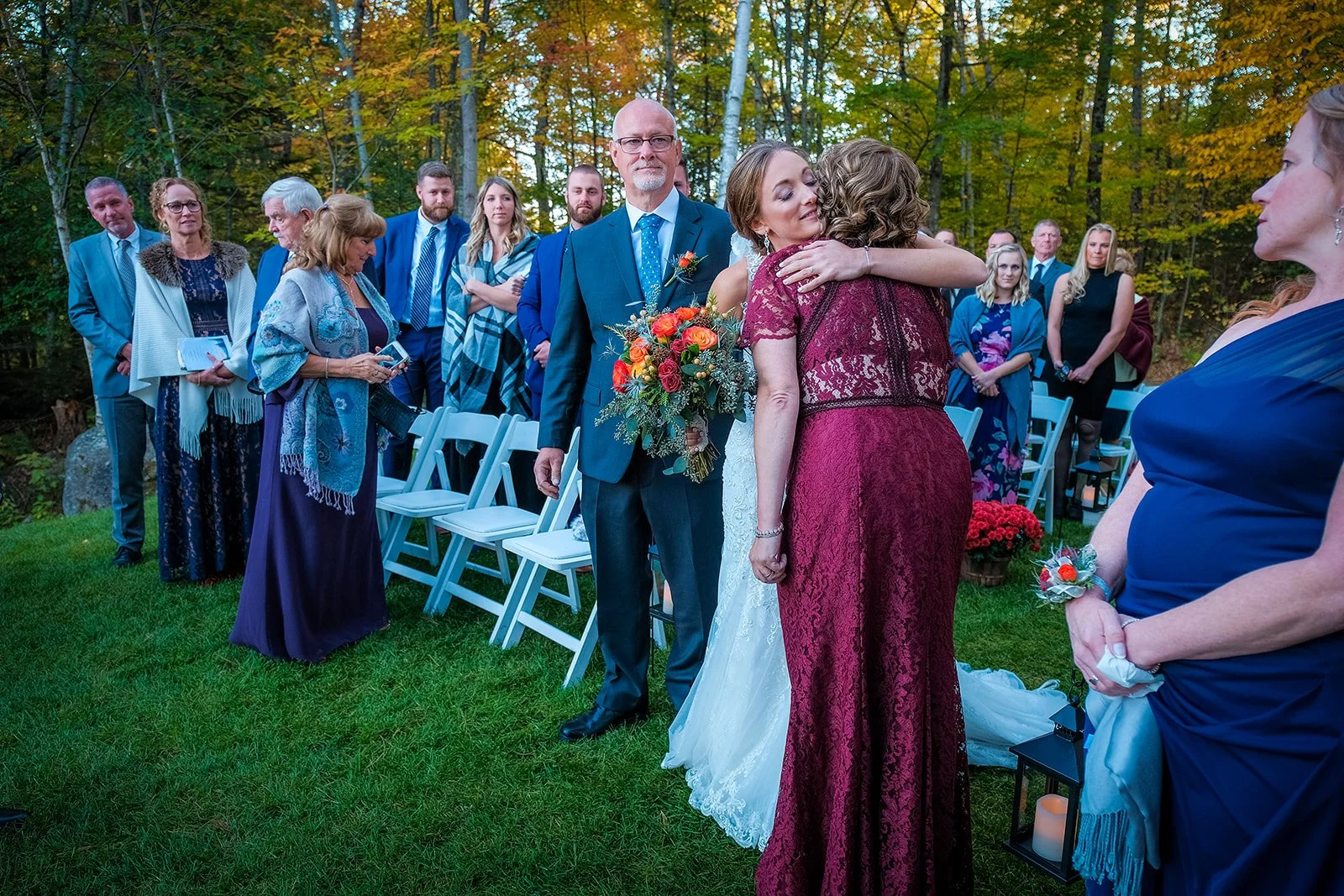 A wedding ceremony outdoors with guests. A bride hugging a woman, with a man holding a bouquet of flowers standing nearby. Guests are dressed formally, some using shawls and holding programs, amidst green grass and trees with fall foliage.