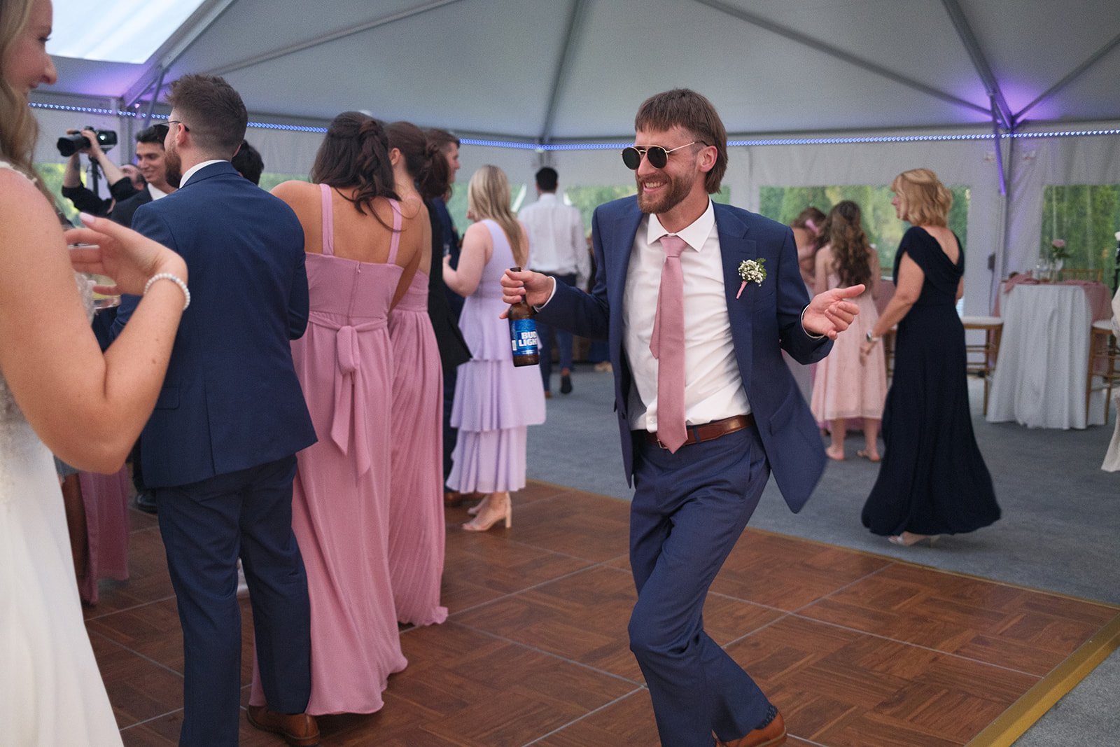 Man in a blue suit with sunglasses dancing at a wedding reception under a tent, surrounded by other guests in formal attire.