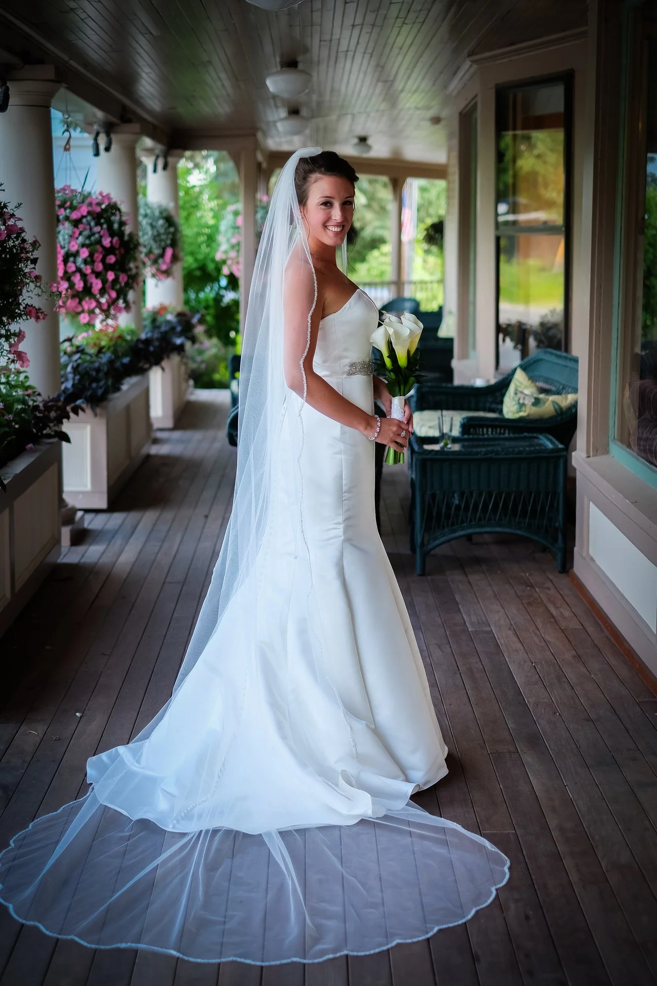 A bride in a white wedding dress with a long veil holding a bouquet of white calla lilies, standing on a wooden porch with hanging pink flowers and greenery in the background.