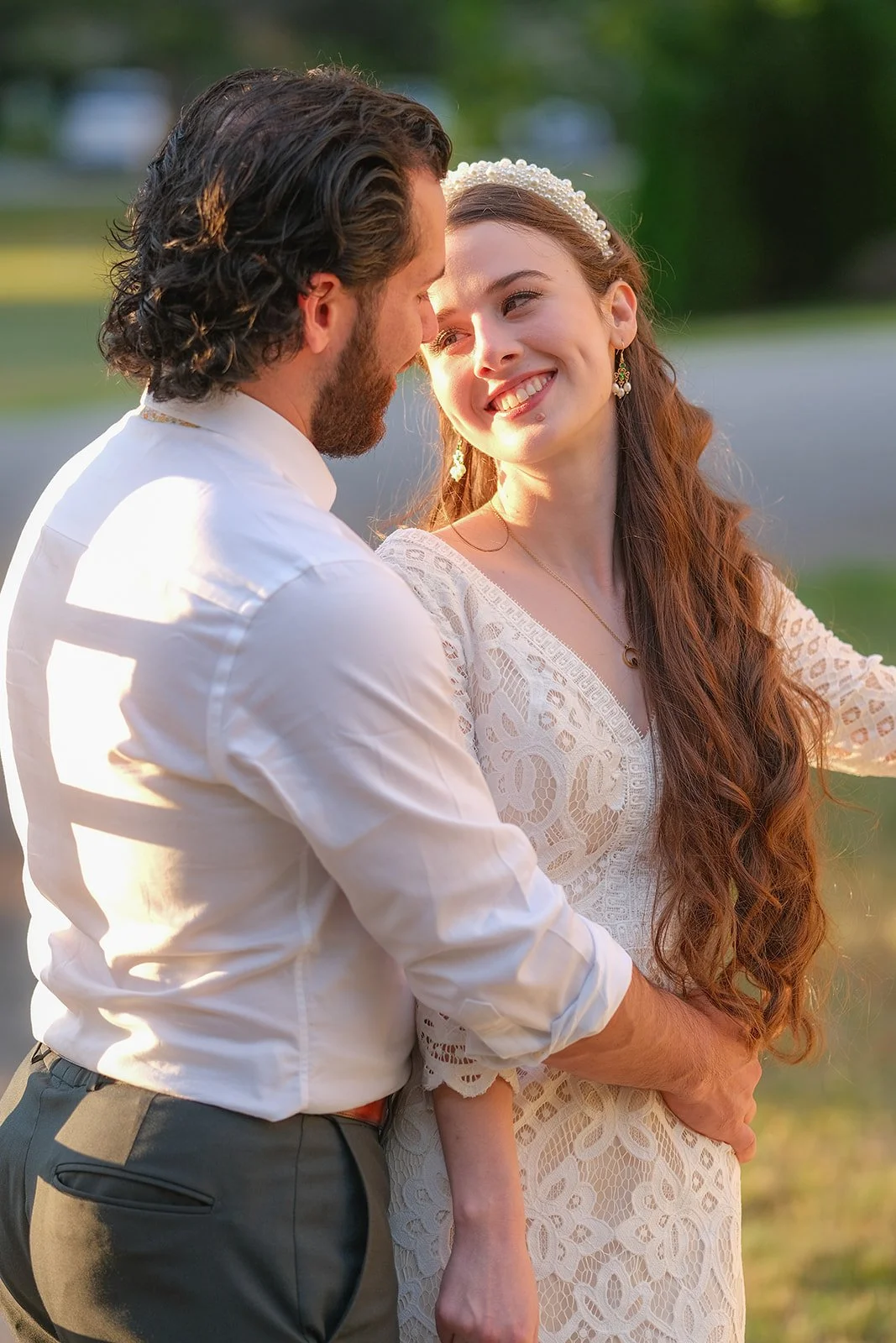 A man and woman stand very close, looking into each other's eyes, in a romantic moment outdoors at sunset. The woman wears a white lace dress and a pearl headband, while the man is dressed in a white shirt and dark trousers.
