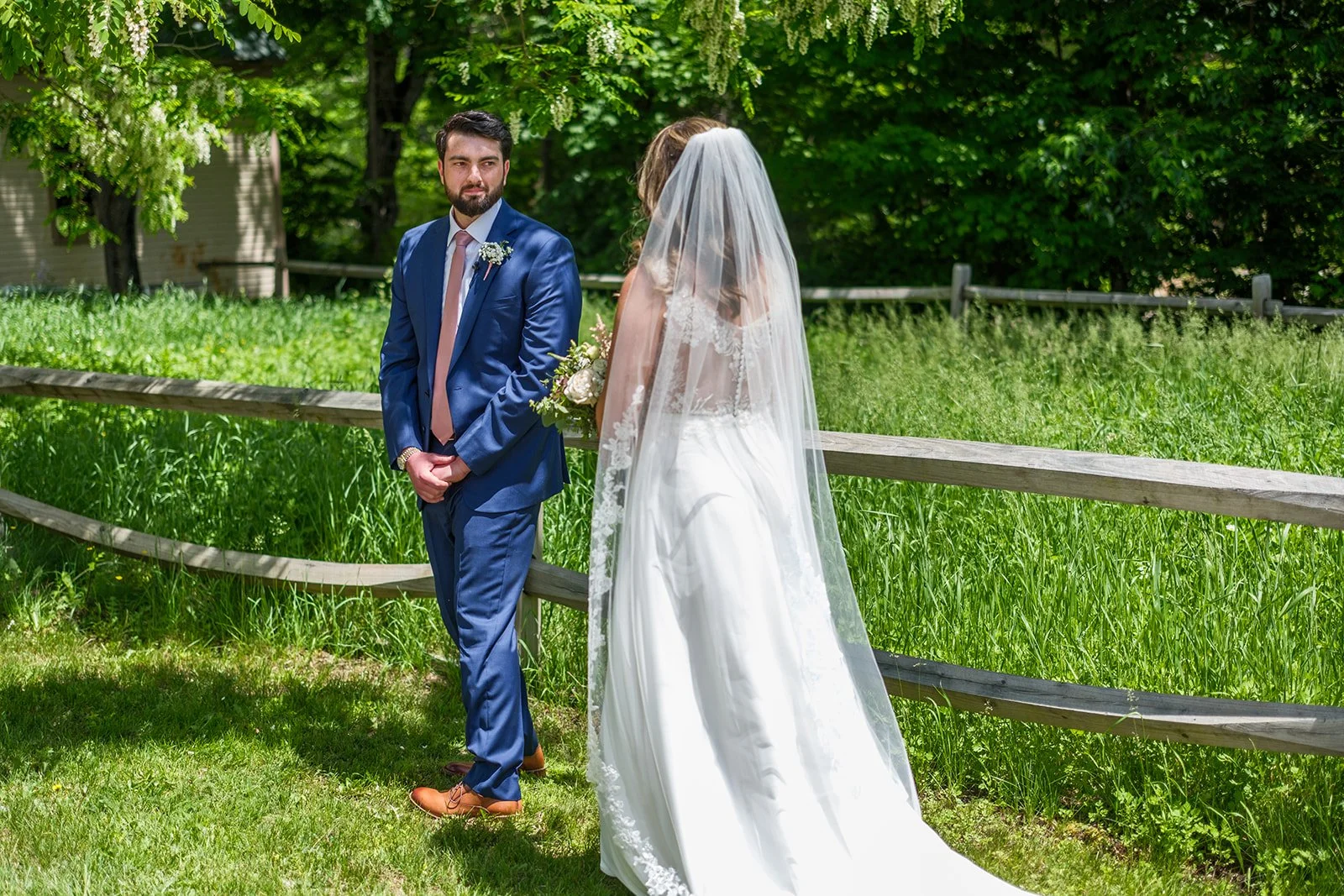 A bride and groom standing outdoors on a sunny day with lush green trees and grass in the background. The groom is wearing a blue suit and the bride is wearing a white wedding dress and veil, holding a bouquet of flowers.