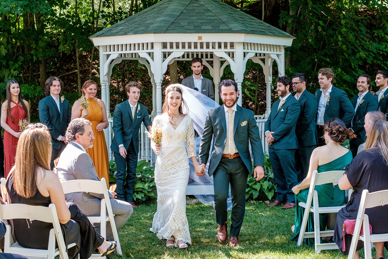 A bride and groom walking hand in hand down the aisle at an outdoor wedding ceremony, surrounded by guests seated on white chairs and wedding party members standing in front of a white gazebo, with green trees in the background.