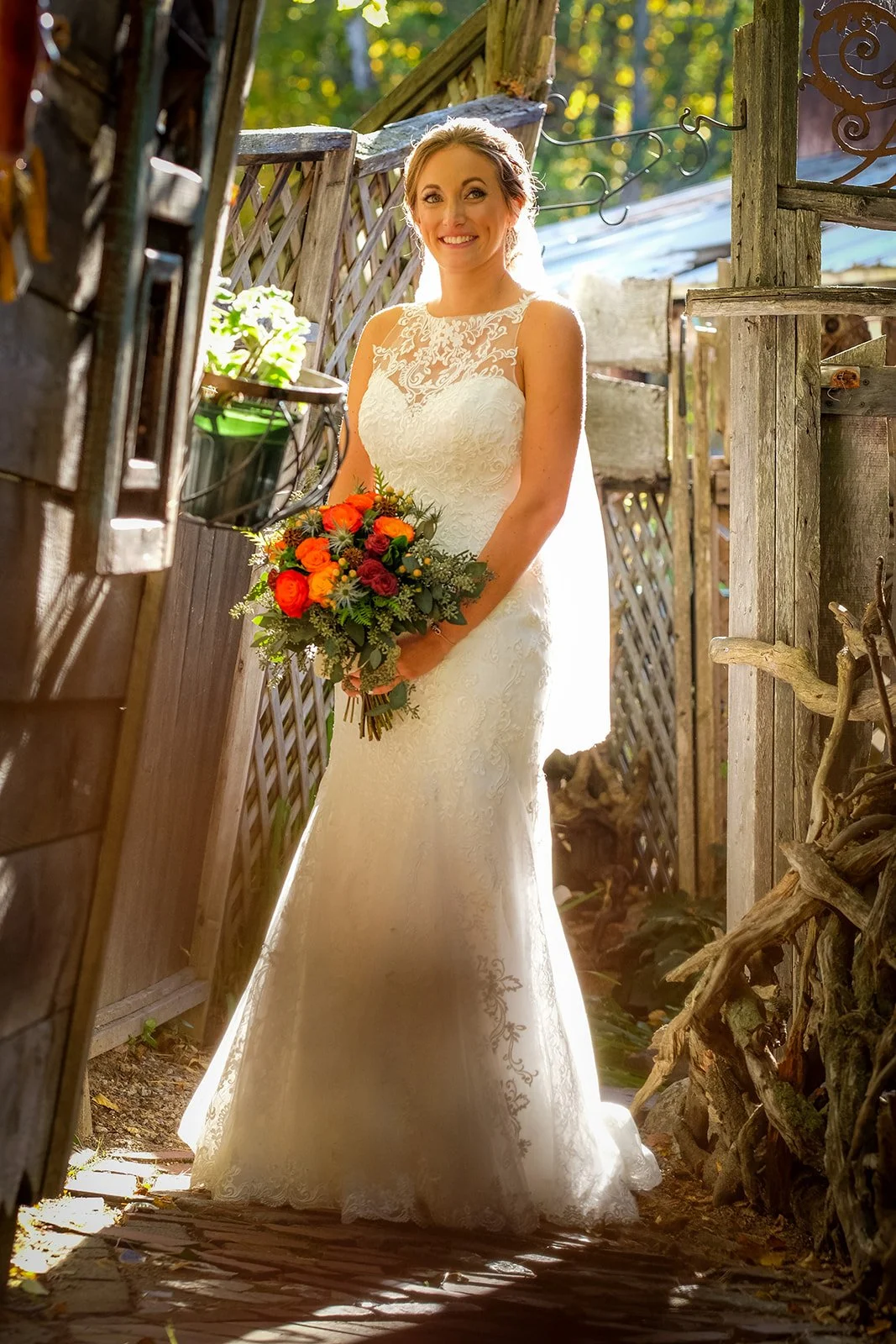 A bride in a white lace wedding dress holding a colorful bouquet, standing in a rustic outdoor setting with sunlight filtering through trees.