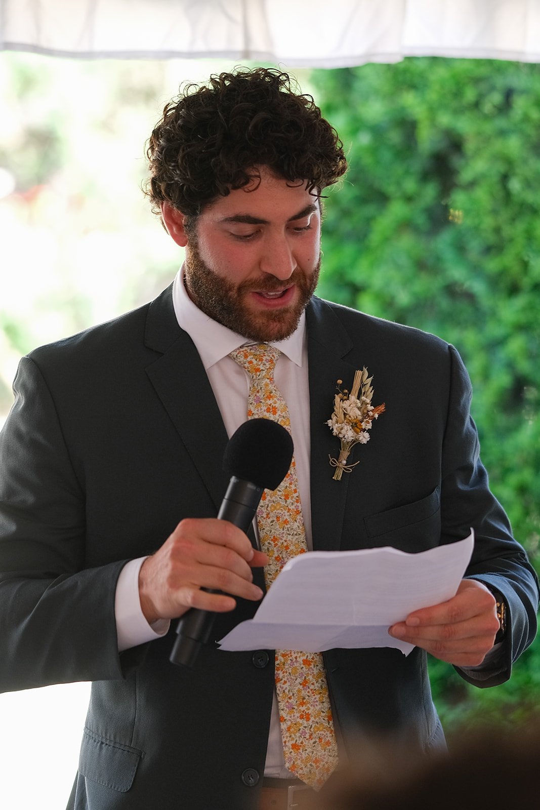 A man with curly hair and a beard, wearing a dark suit and a floral tie, is holding a microphone and reading from a piece of paper during a formal event with greenery in the background.