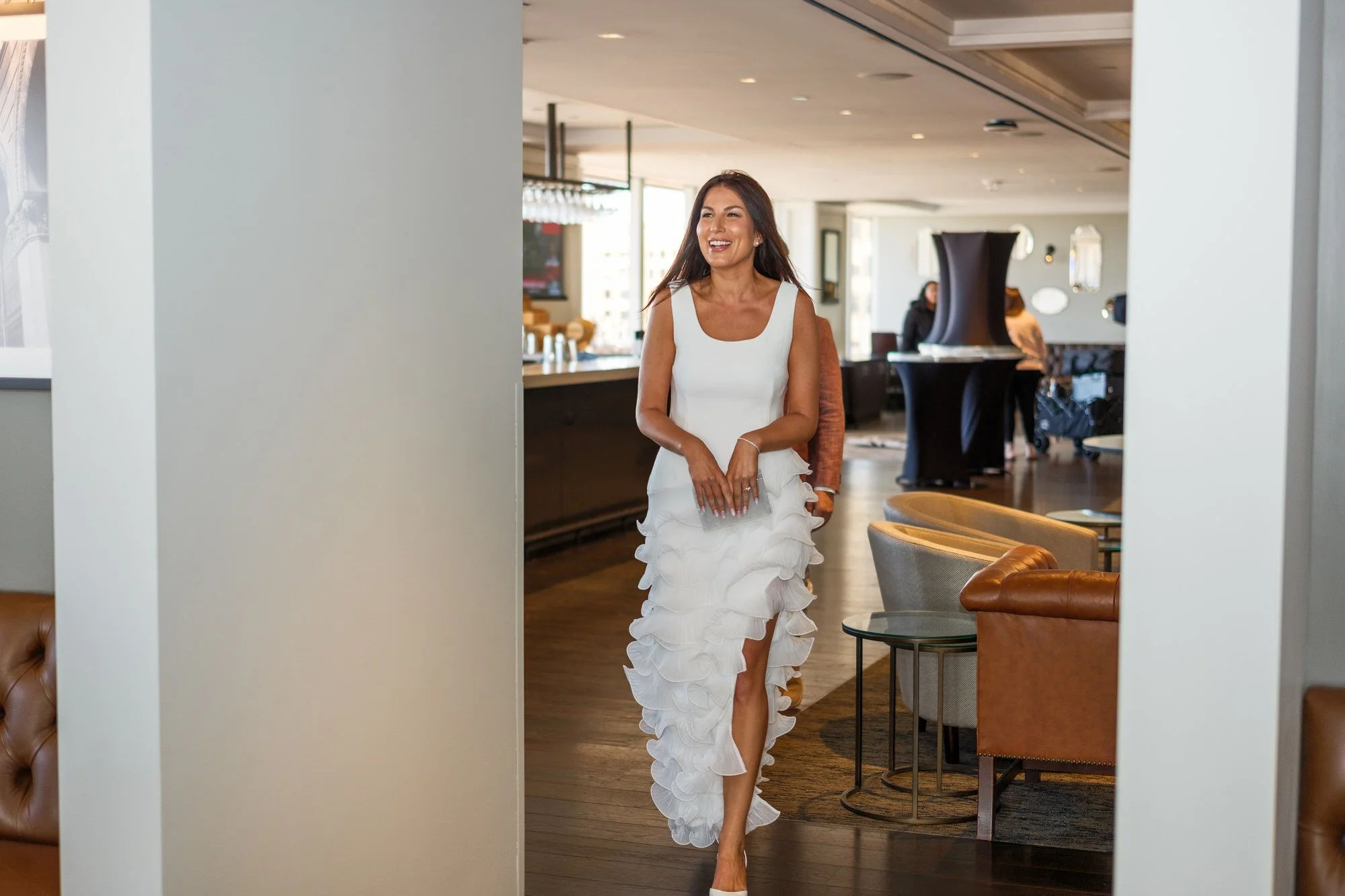 Bride Lauren walking through the elegant lounge at the UMass Club in Boston wearing a white tiered ruffle dress, smiling and relaxed the evening before her wedding