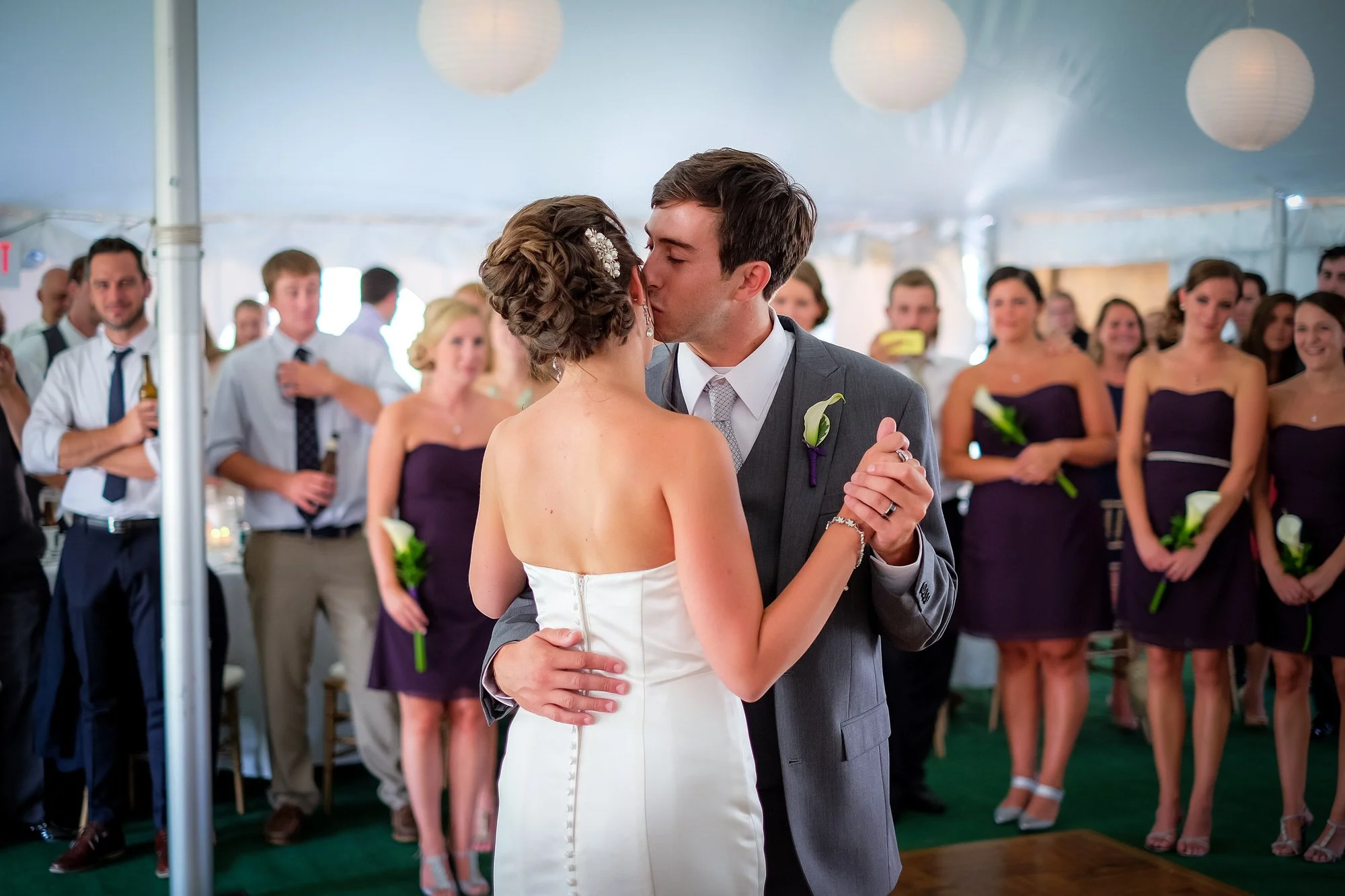 A bride and groom share a dance during their wedding reception, surrounded by friends in formal attire inside a decorated tent.