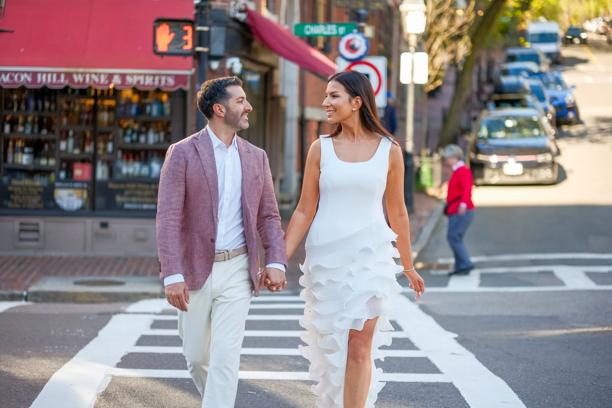Lauren and Charlie walking hand-in-hand across a crosswalk on Charles Street in Beacon Hill, smiling at each other with the Beacon Hill Wine and Spirits shop visible behind them