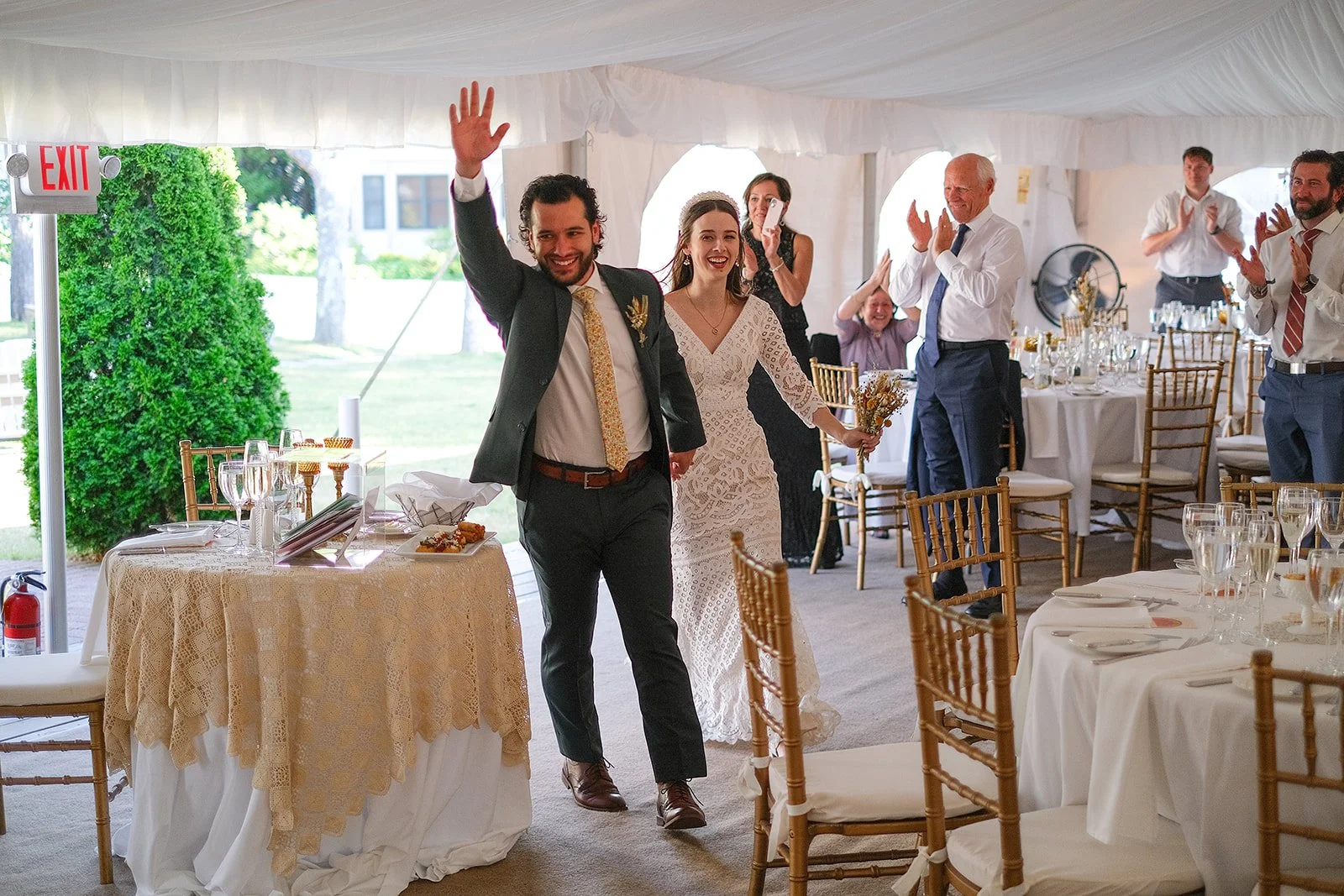 A wedding reception inside a tent with a bride and groom walking in, celebrating, with guests clapping and smiling.