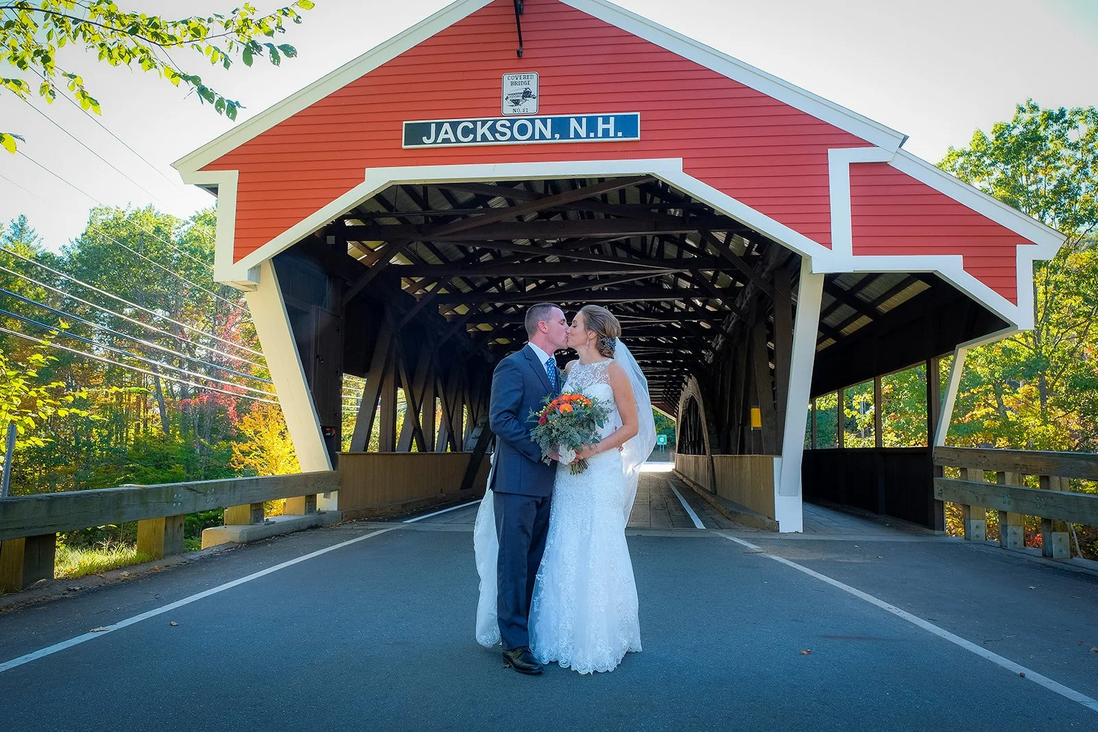 A newlywed couple sharing a kiss under a red covered bridge in Jackson, New Hampshire. The bride holds a bouquet of flowers, and the groom is dressed in a blue suit. The scene is outdoors with trees in the background.