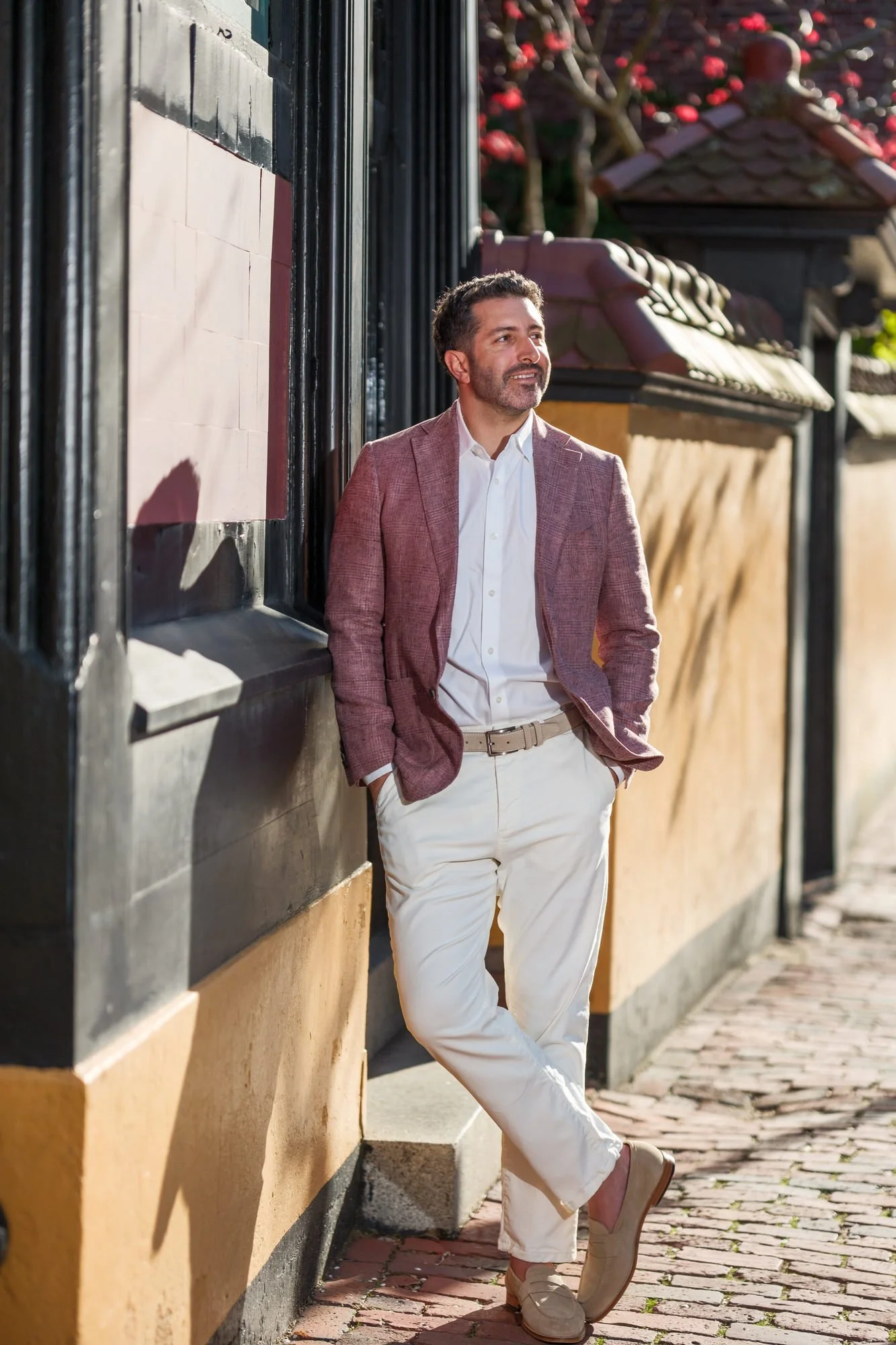 Groom Charlie leaning casually against a colorful building facade on a cobblestone street in Beacon Hill, Boston, in a blush linen blazer, gazing confidently off-camera