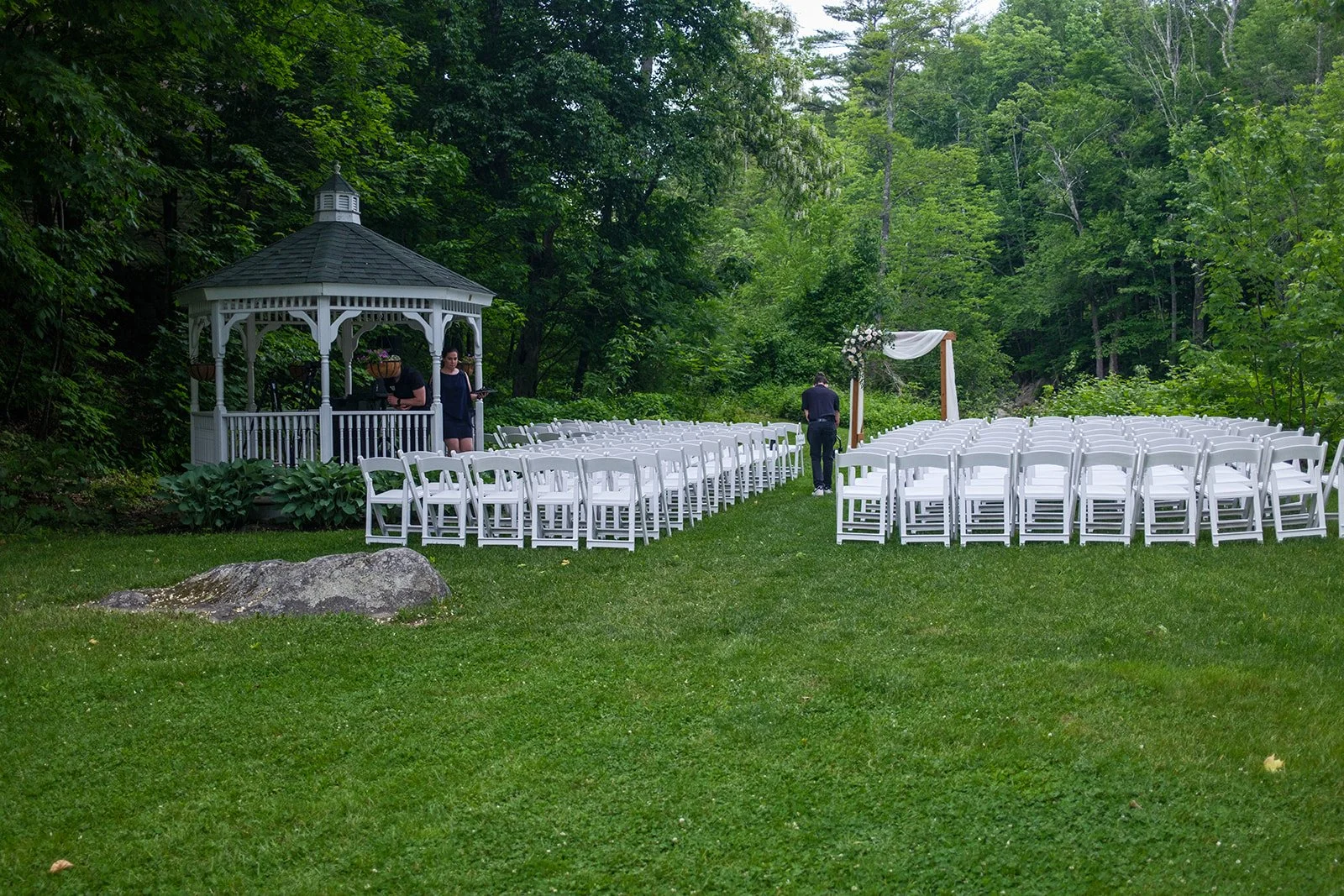 Wedding ceremony setup with white chairs arranged in two sections facing an arch with white fabric, in a lush green outdoor setting with trees and a small white gazebo.