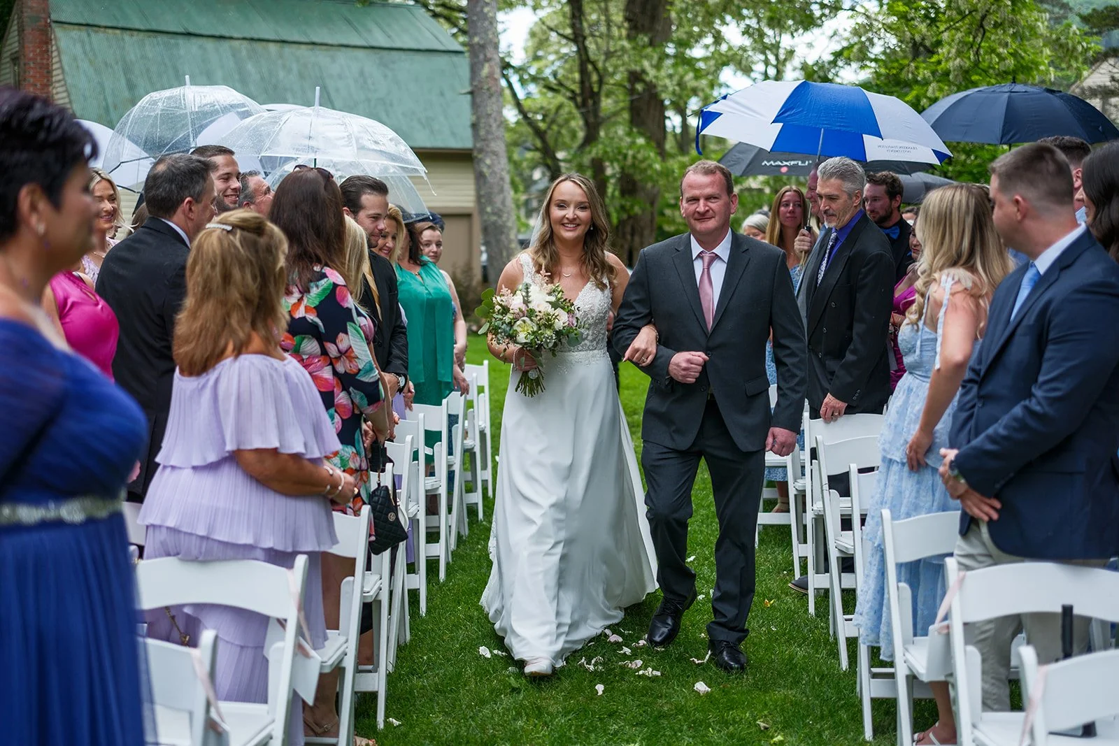 A bride in a white wedding dress holding a bouquet, walking down the aisle with a man in a gray suit, during an outdoor wedding ceremony on a rainy day. Guests are standing under umbrellas, watching and smiling.