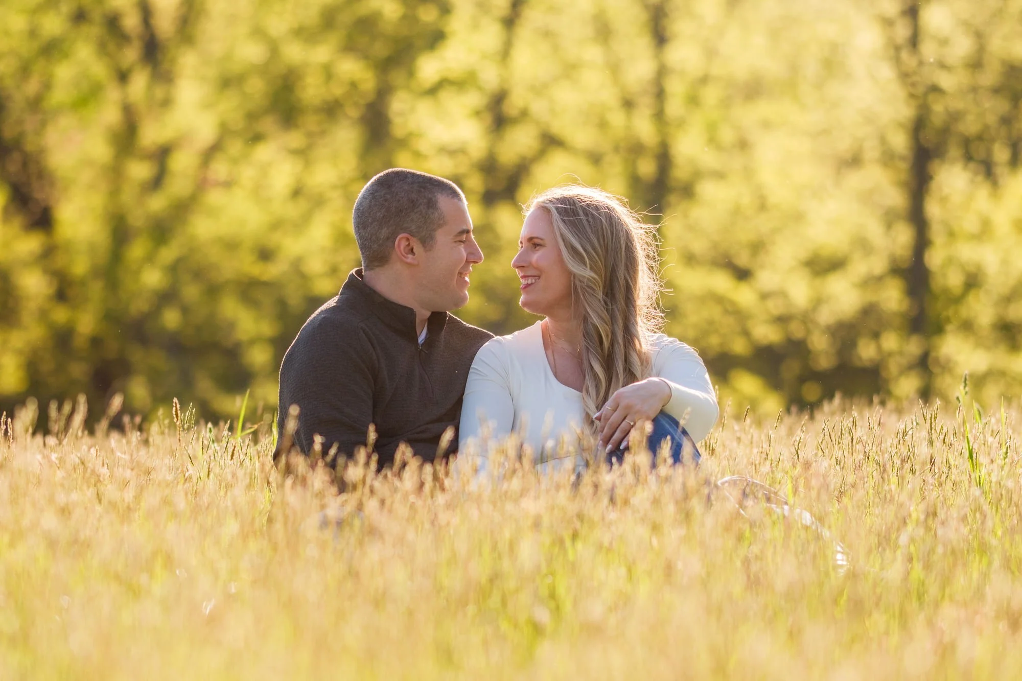 Engaged couple sitting close together in tall golden grass smiling at each other in warm afternoon light at Maudslay State Park