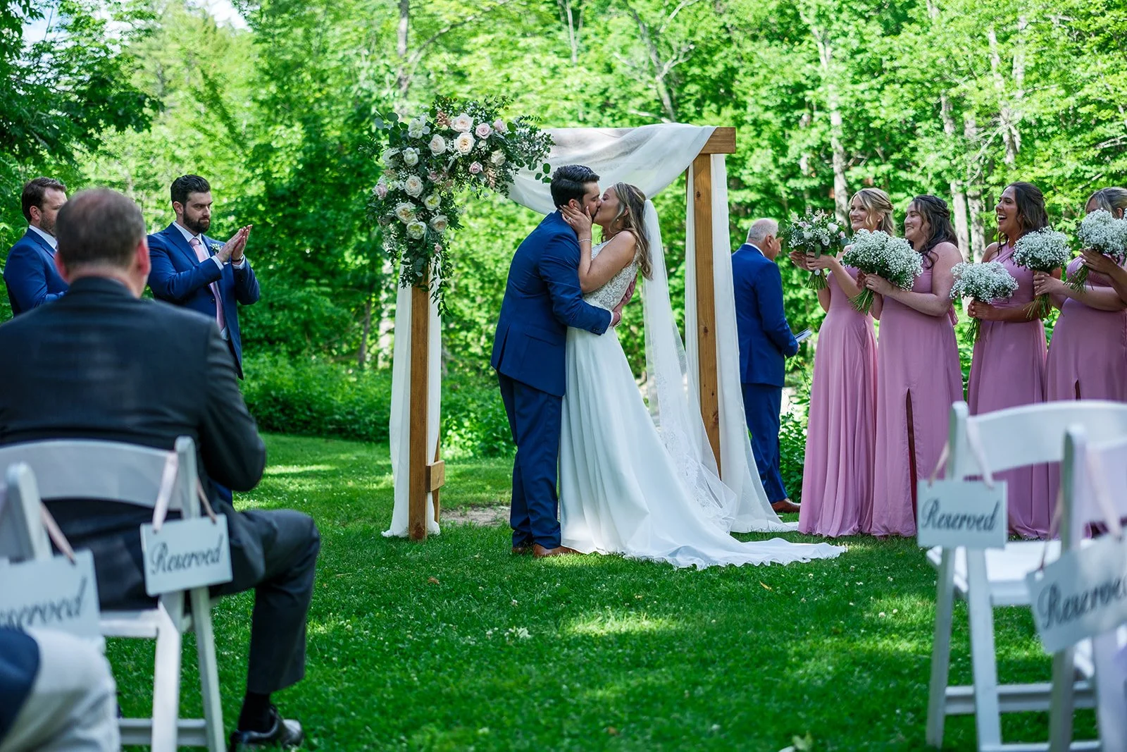 A couple getting married kissing under a wooden arch decorated with white and pink flowers, surrounded by bridesmaids in pink dresses holding bouquets and groomsmen in blue suits, in a lush outdoor garden setting.