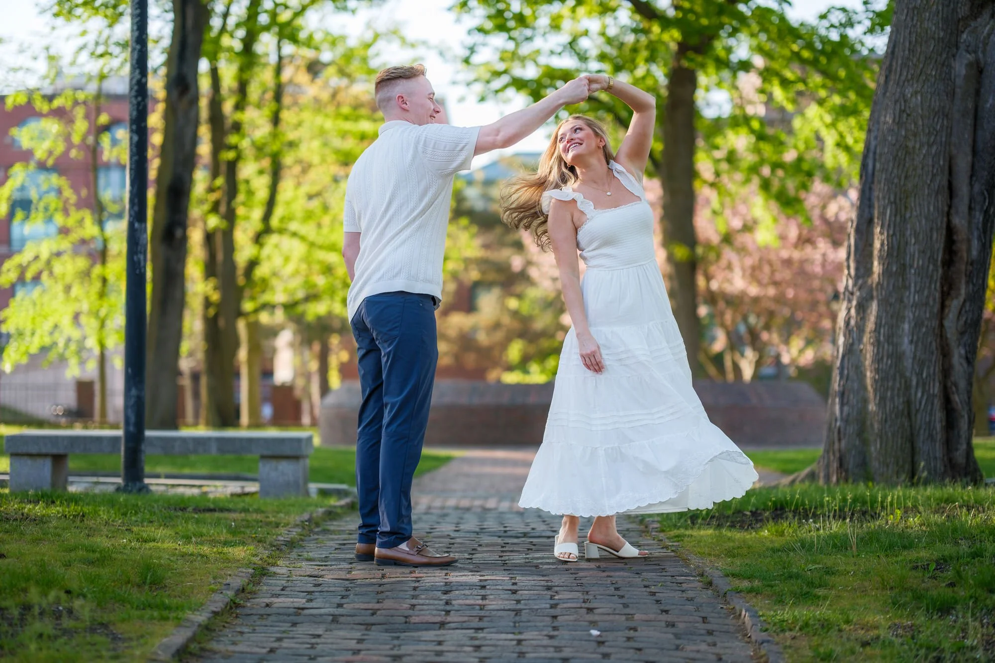 Connor spins Melanie in a joyful dance on a brick park path surrounded by lush spring greenery at Prescott Park, Portsmouth, NH