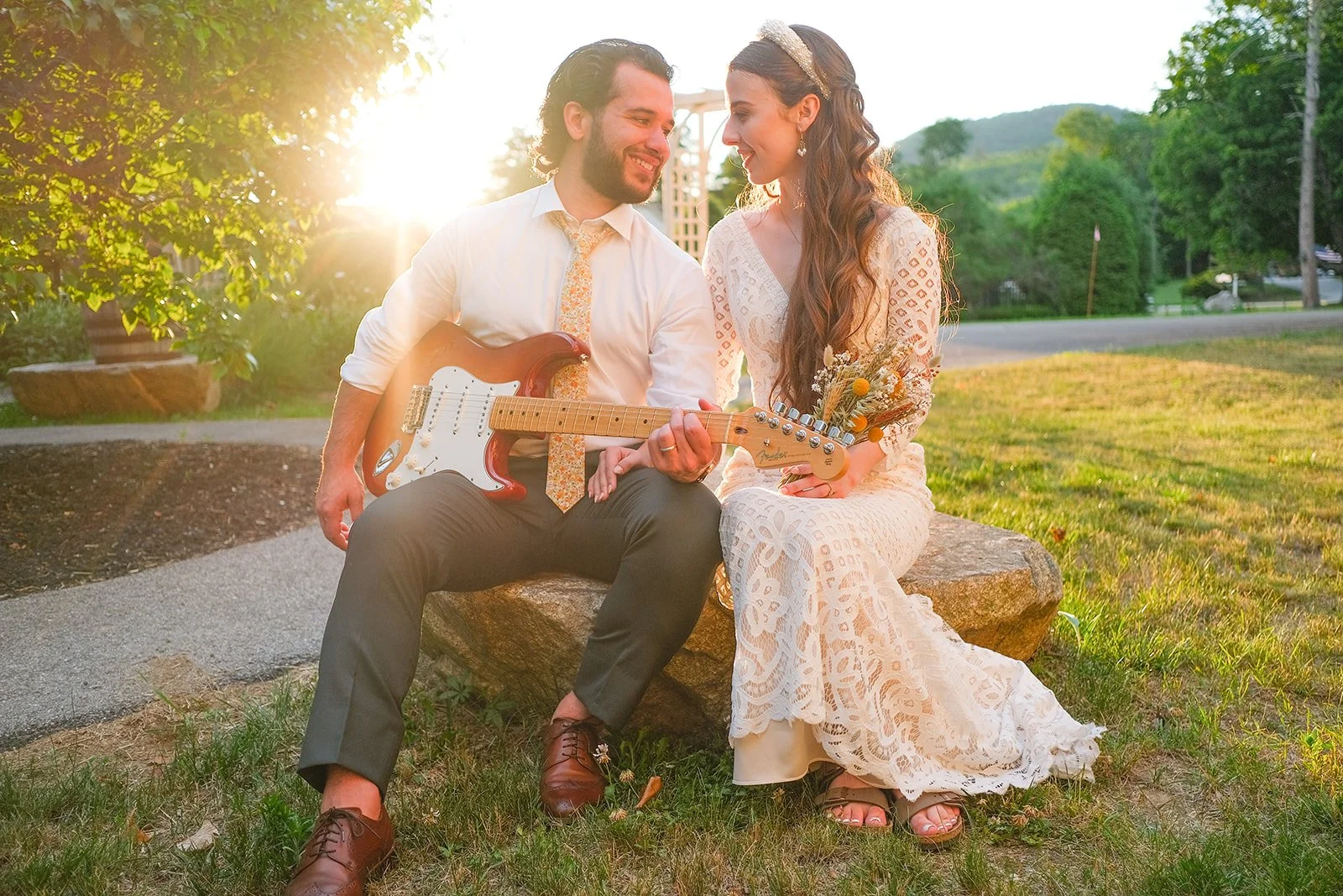 A couple sitting on a rock bench outdoors during sunset, with the man playing an electric guitar and the woman holding a bouquet of flowers. They are dressed in vintage-style clothing, and the scene is surrounded by green trees and grass.