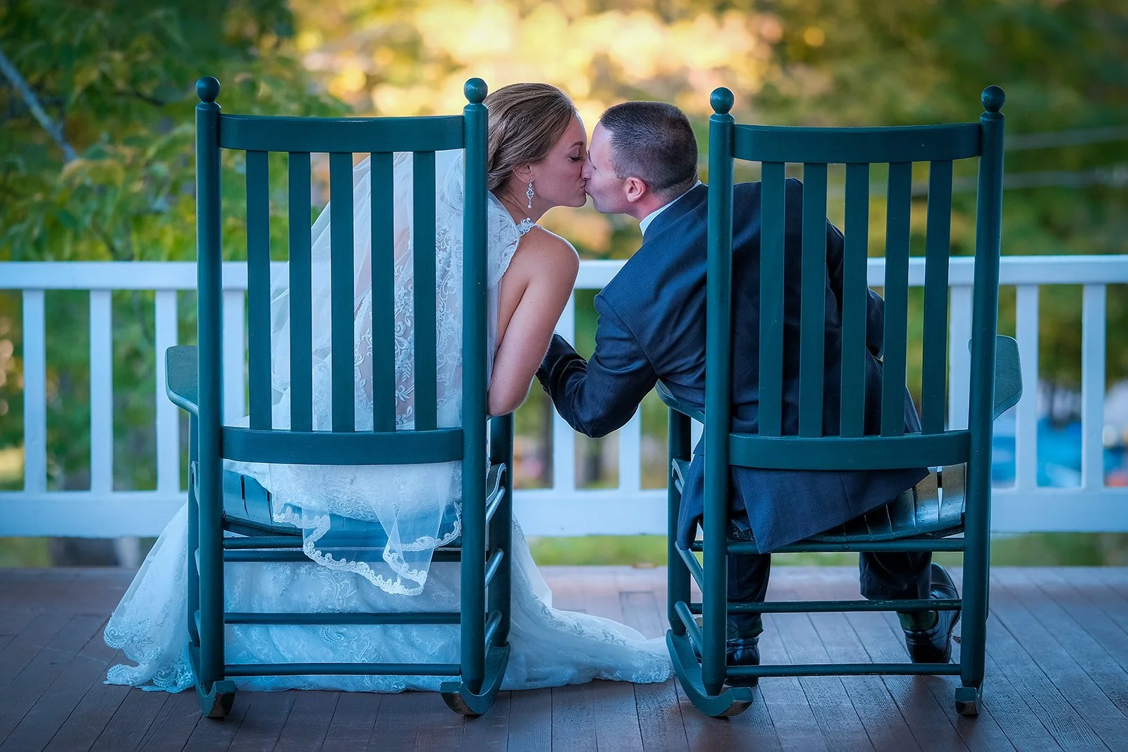 A bride and groom sitting on a porch, kissing each other through two blue rocking chairs with a white railing and trees in the background.