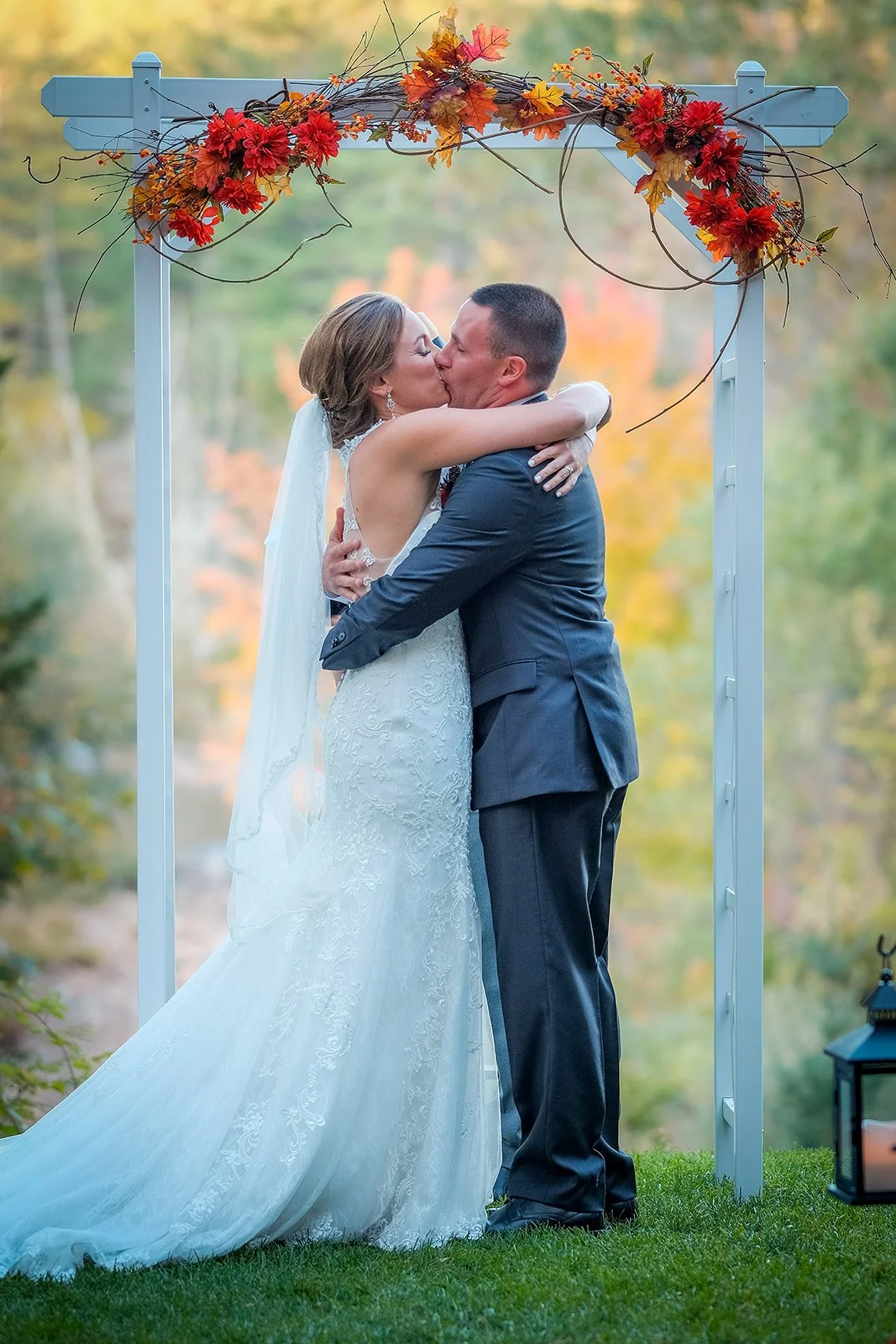 Bride and groom kissing under a decorated outdoor arch during a wedding ceremony with autumn foliage in the background.