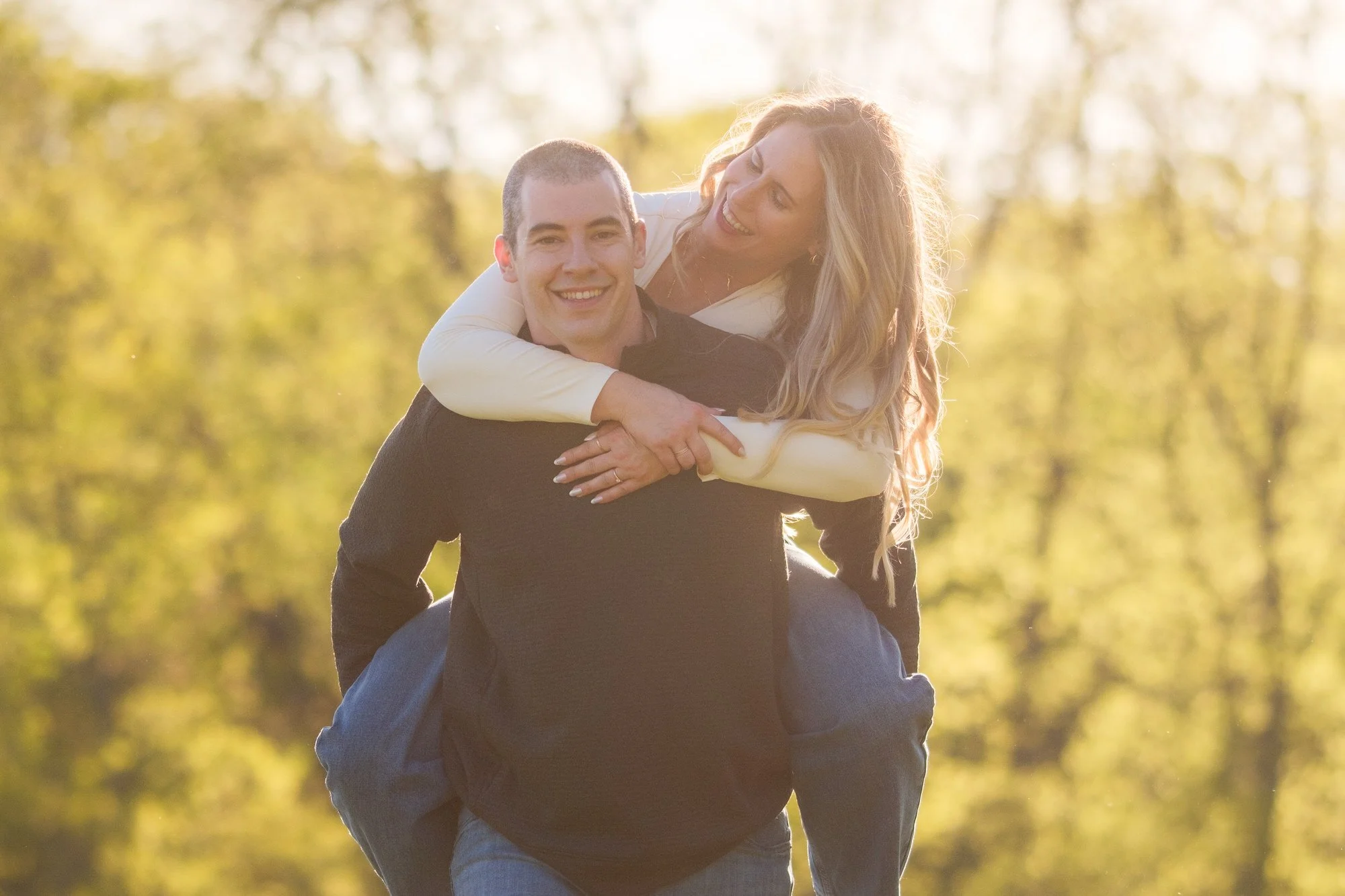 Bride-to-be riding piggyback on her laughing fiancé in glowing golden hour light at Maudslay State Park