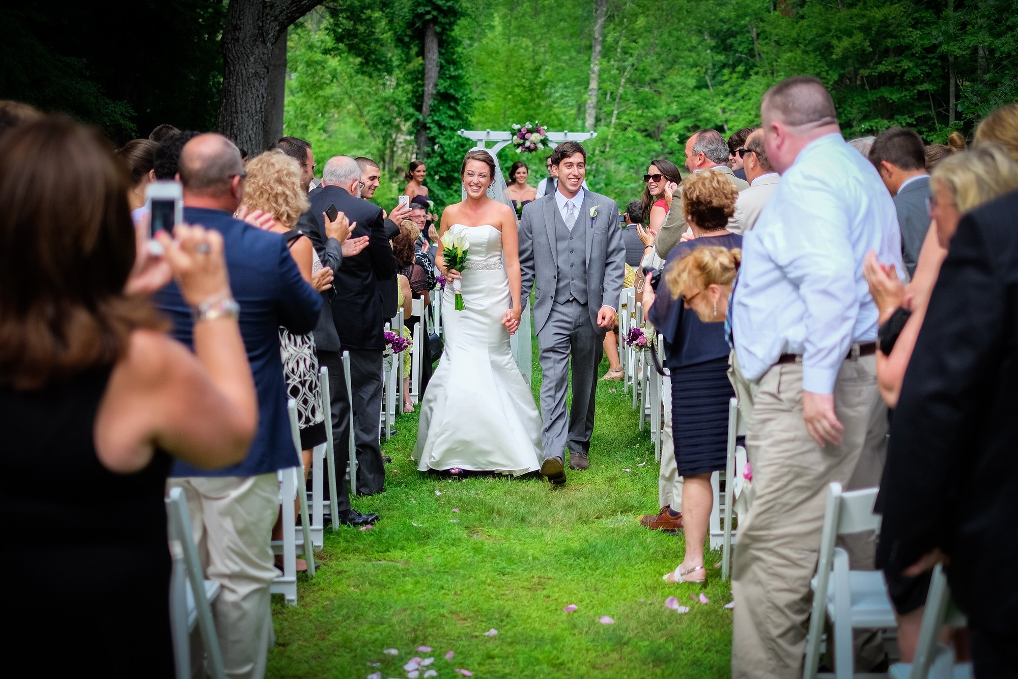 A bride and groom walking down the aisle holding hands, surrounded by smiling guests at an outdoor wedding ceremony in a green, wooded area.