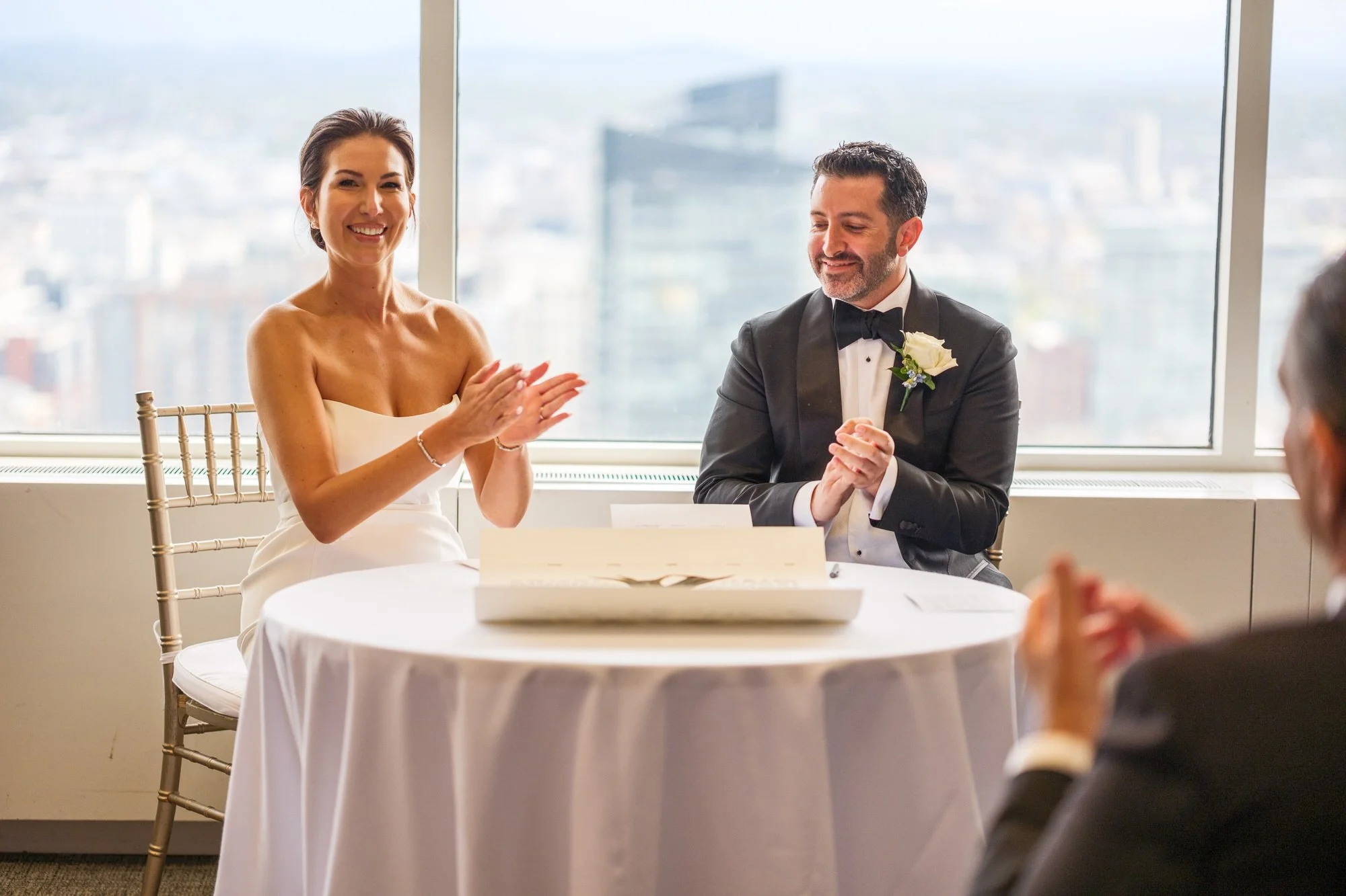 Bride and groom smiling and applauding during their ketubah signing ceremony at the UMass Club Boston with city views