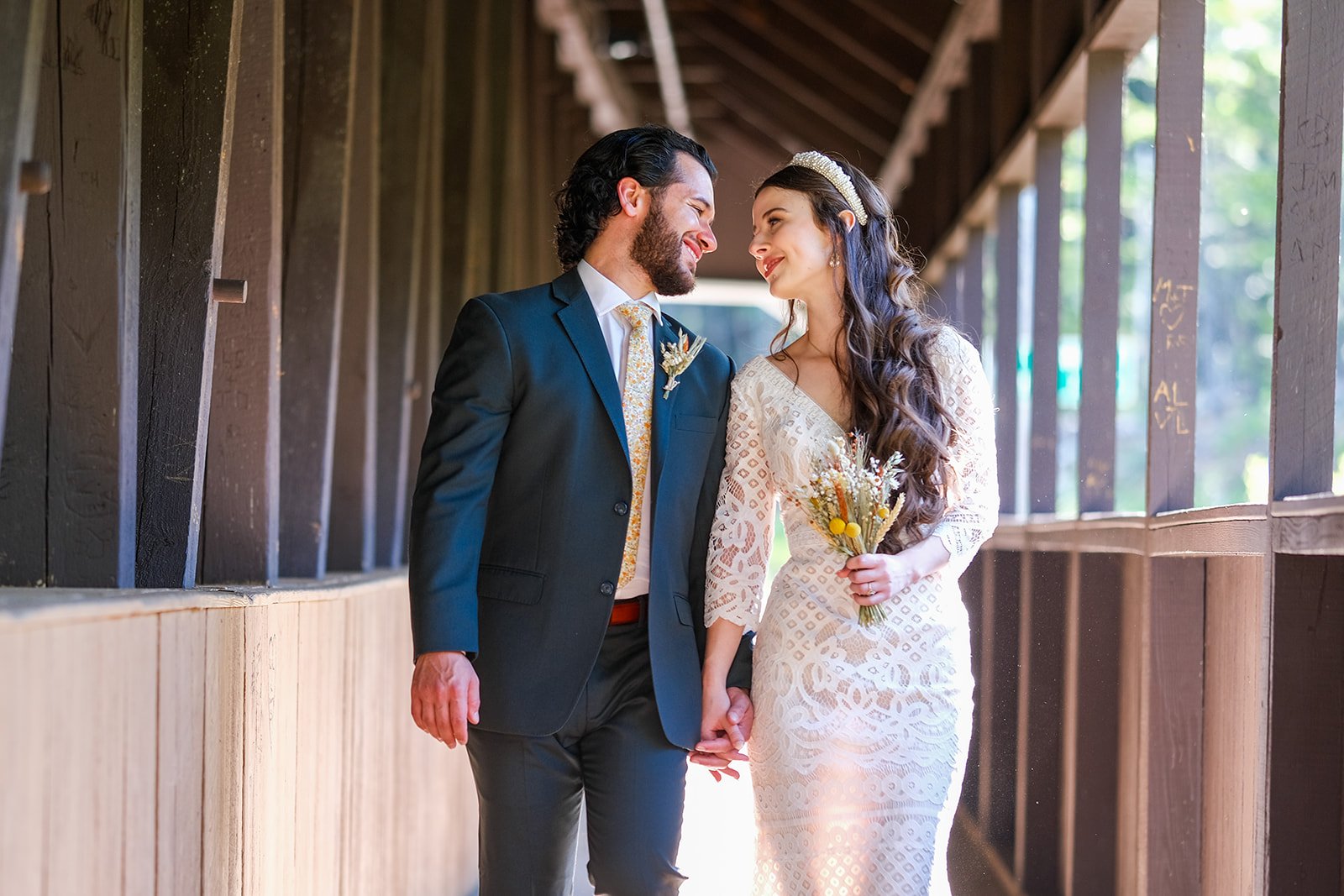 A bride and groom holding hands and looking at each other, standing in a wooden corridor. The bride is wearing a white lace wedding dress and holding a small bouquet of flowers, while the groom is dressed in a dark suit with a floral tie.