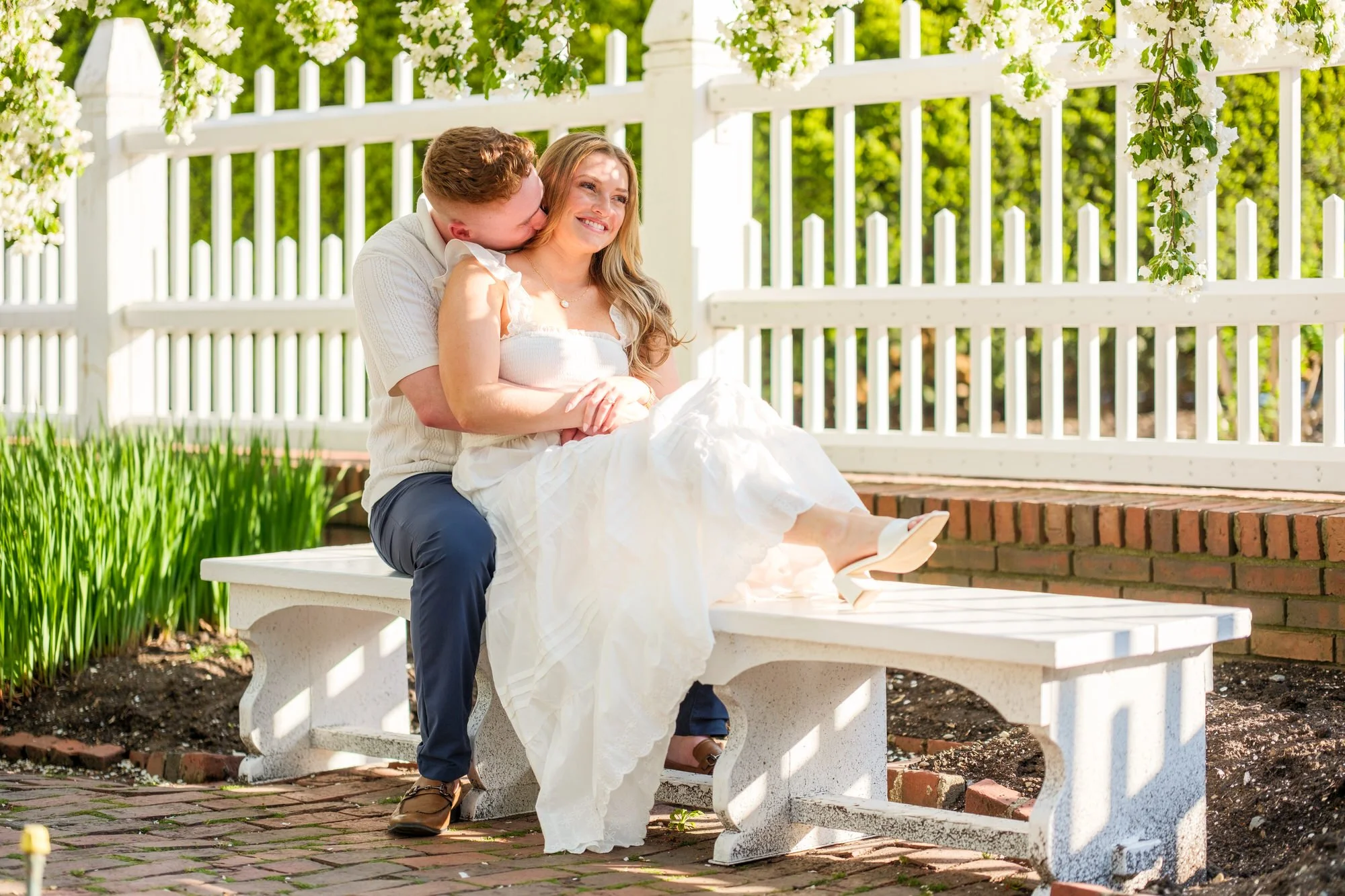 Connor nuzzles into Melanie's neck from behind as she smiles on a white garden bench surrounded by spring blooms at Prescott Park, Portsmouth, NH
