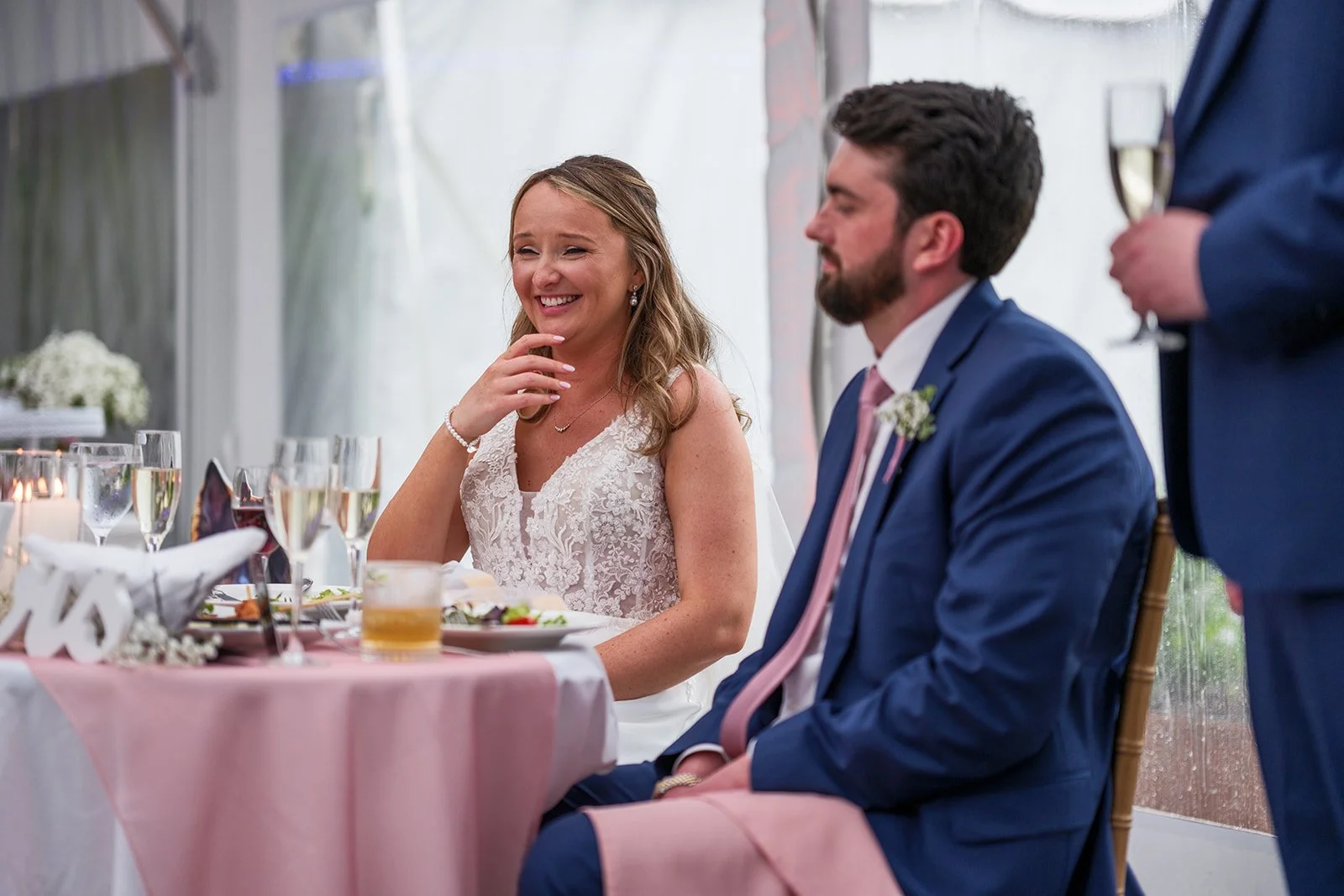 A bride and groom sit at a wedding reception table, smiling. The bride wears a white lace dress, and the groom wears a blue suit with a pink tie. They are surrounded by champagne glasses and floral centerpieces.