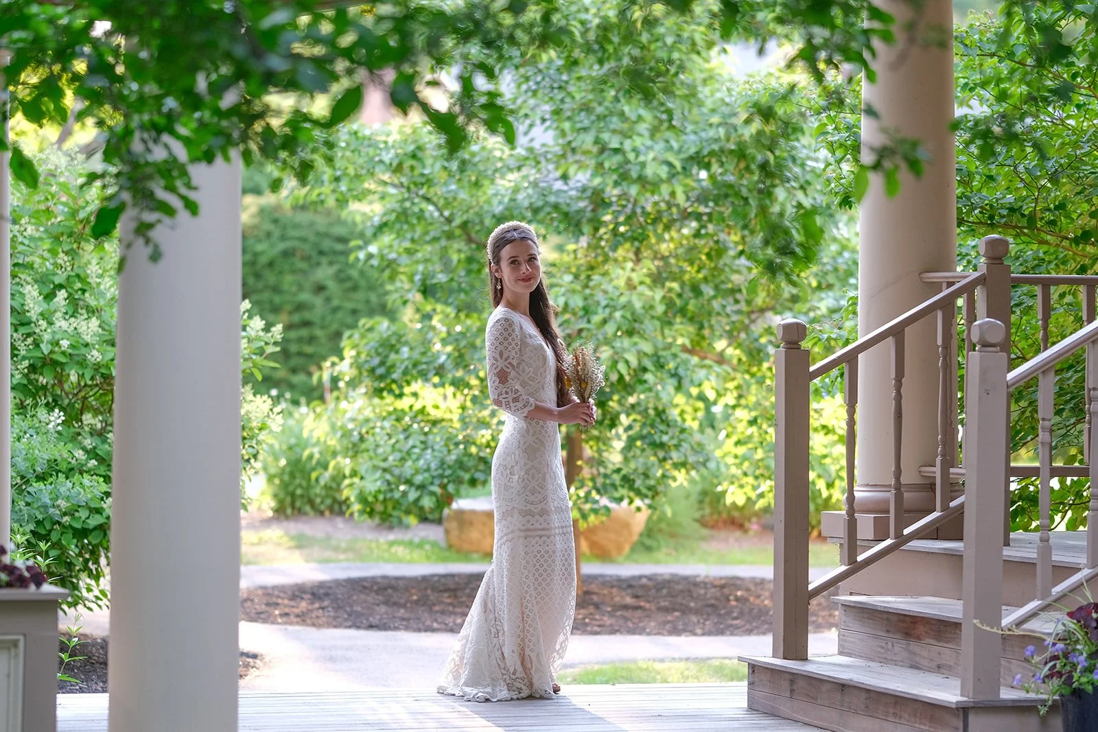 A woman in a white lace dress holding a bouquet standing on a porch surrounded by lush green trees and plants during daytime.