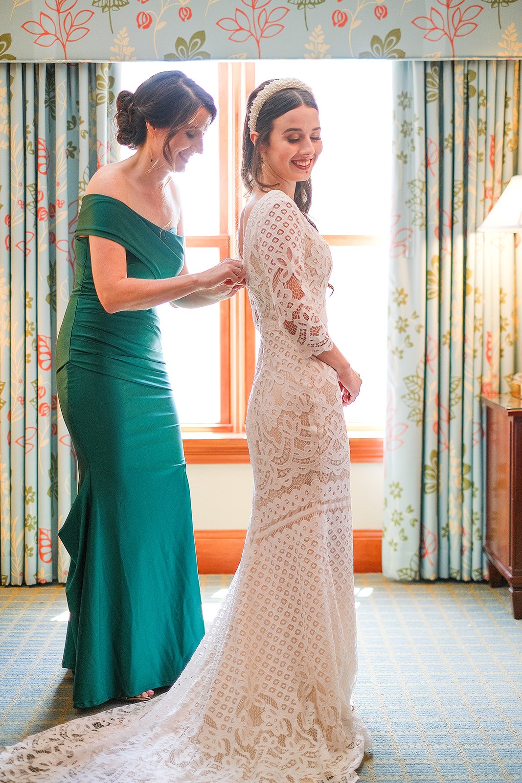 A woman in a white lace wedding dress is smiling while another woman in a green dress helps her with her gown in a brightly lit room with floral curtains.
